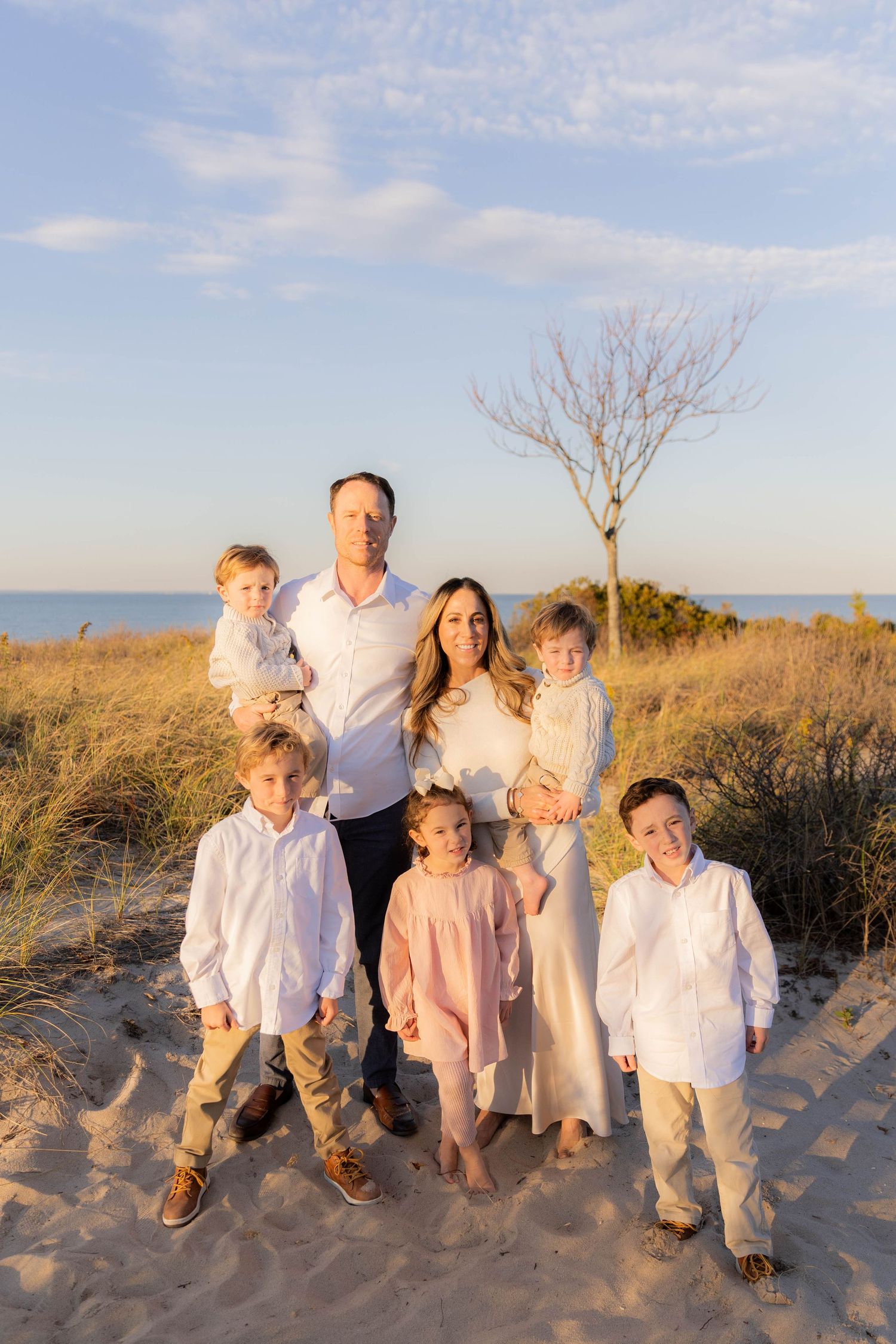 Family wearing white and pink outfits posing together on sandy beach at sunset.