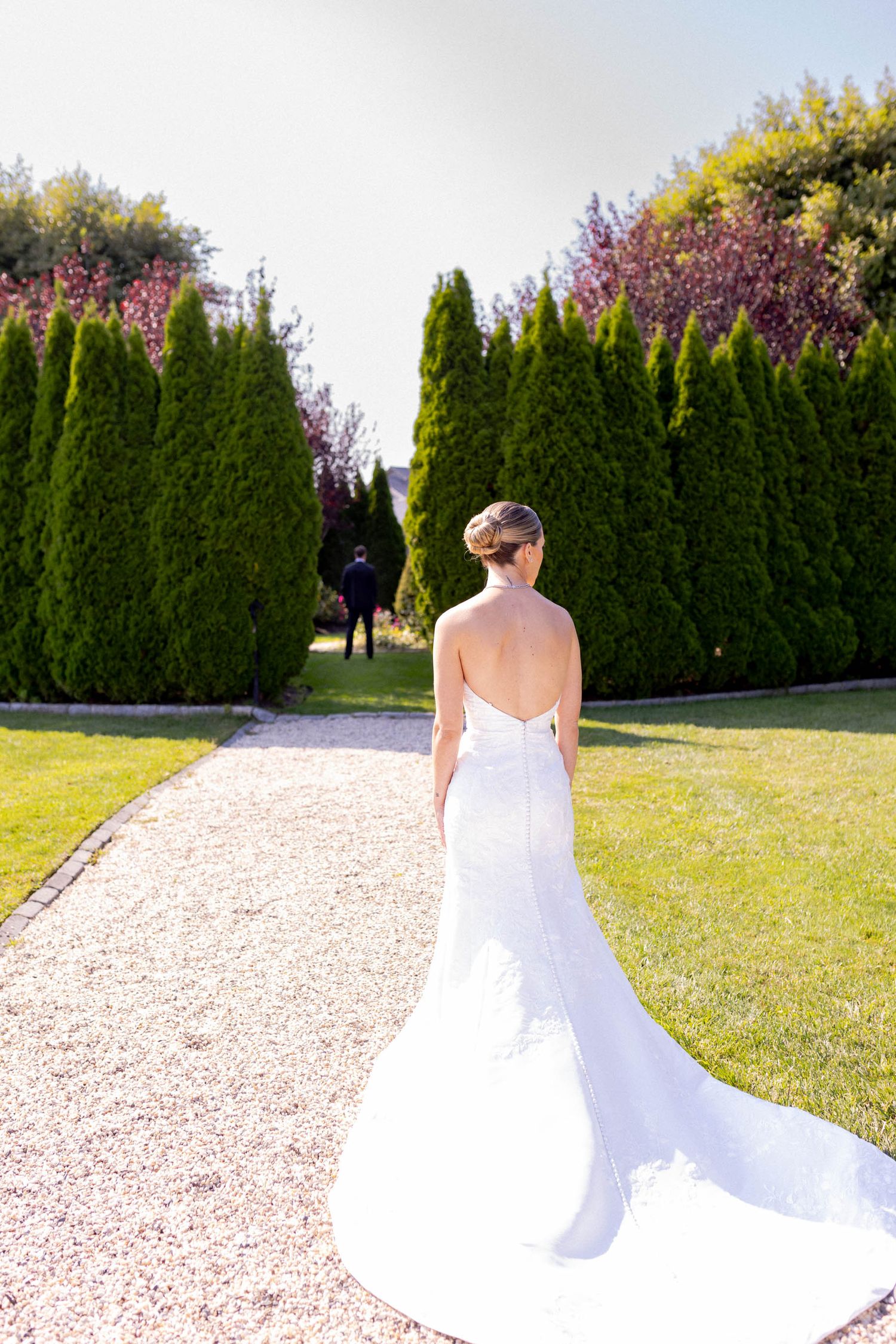 Bride in flowing white wedding dress walks along garden path with tall evergreen hedges and purple flowers.