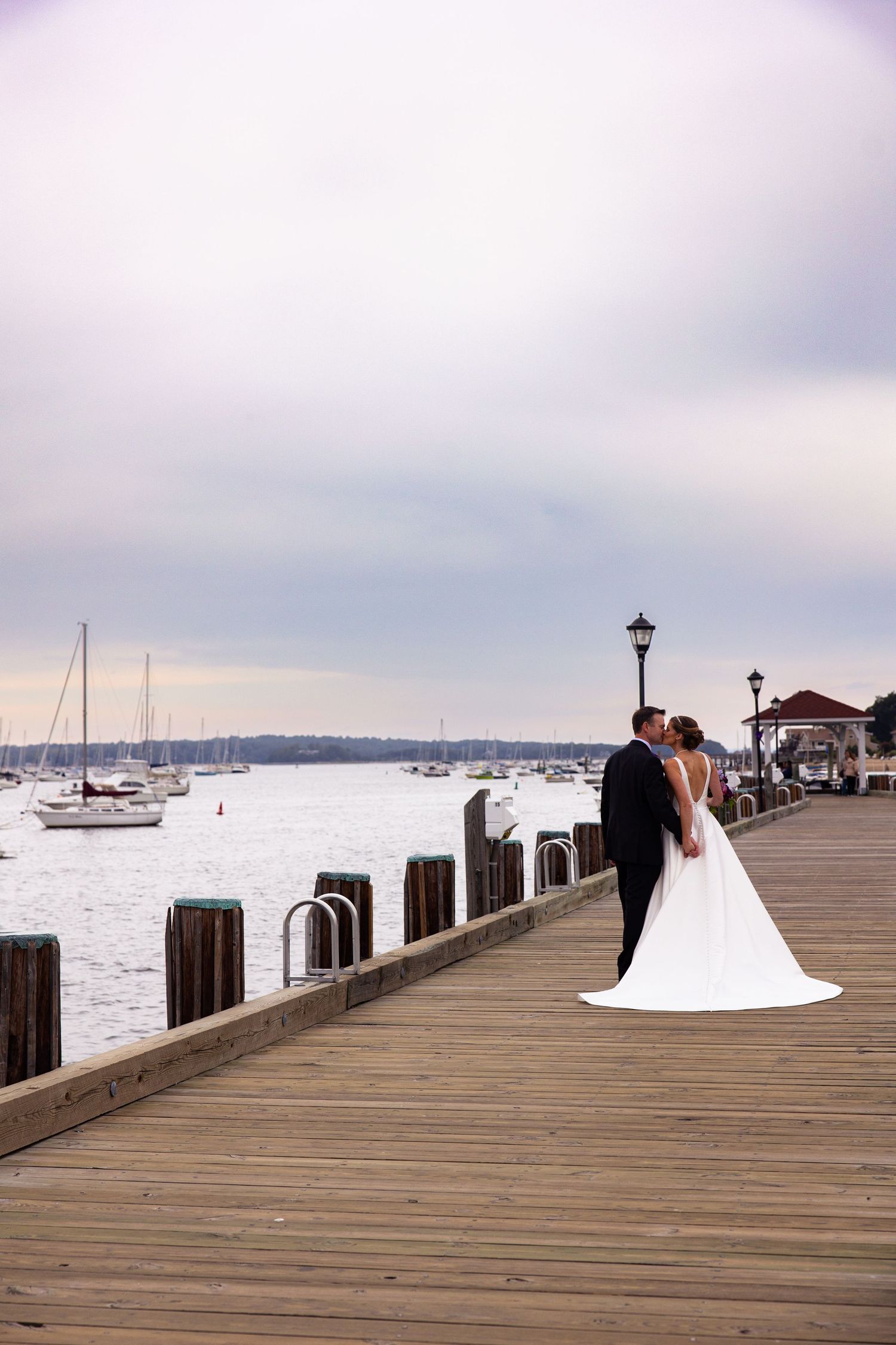 A couple shares a romantic moment on a wooden pier overlooking a harbor with boats in the background.