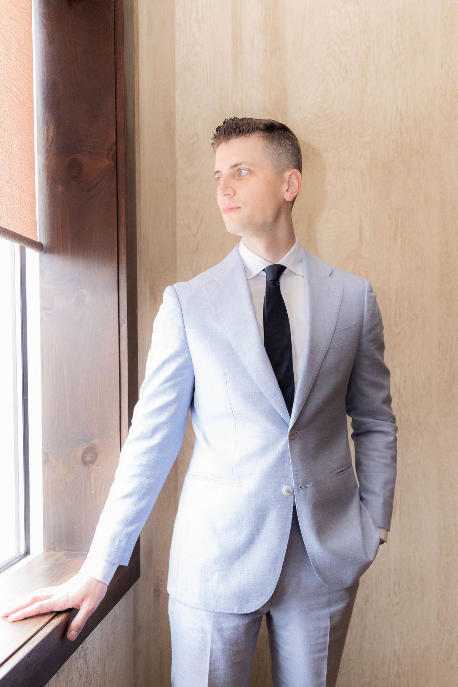A figure wearing a light gray suit jacket and black tie poses against a beige wall near a window.