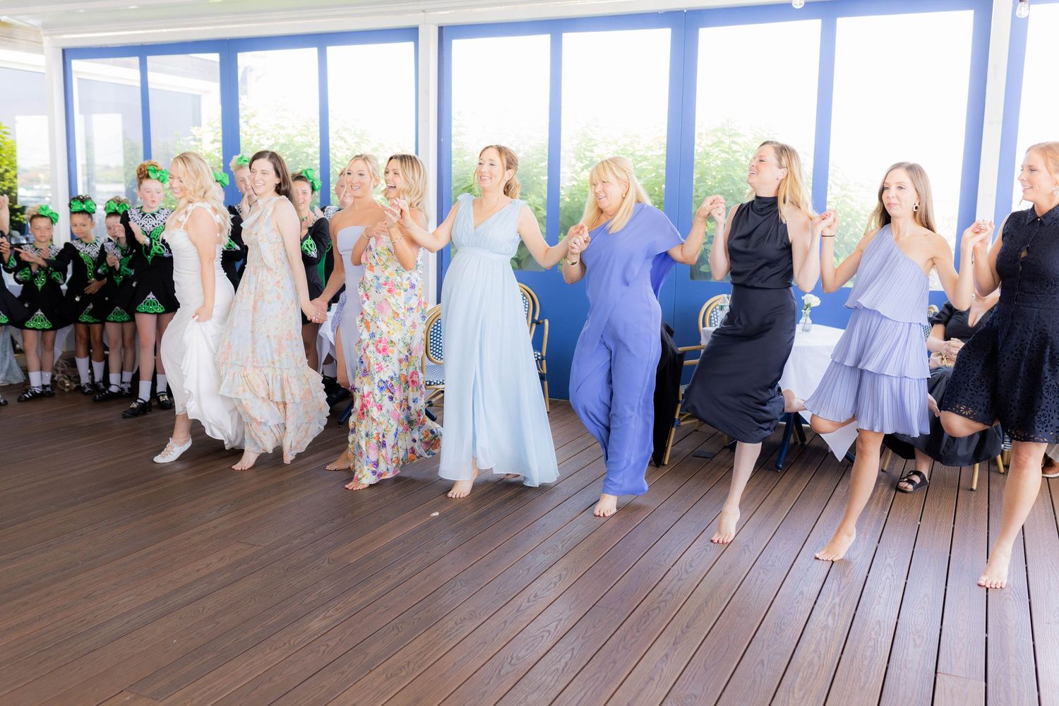 A group of wedding guests dancing joyfully in a bright room with floor-to-ceiling windows and wooden floors.