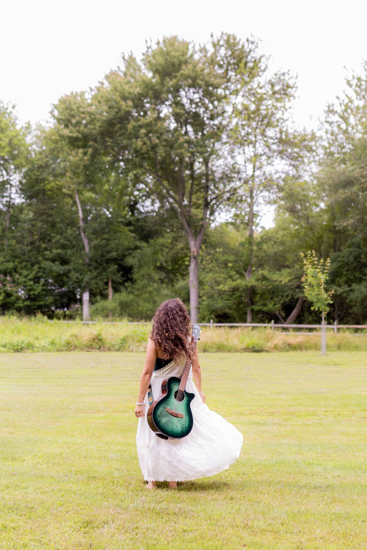 Figure in flowing white dress walks through field carrying turquoise acoustic guitar.