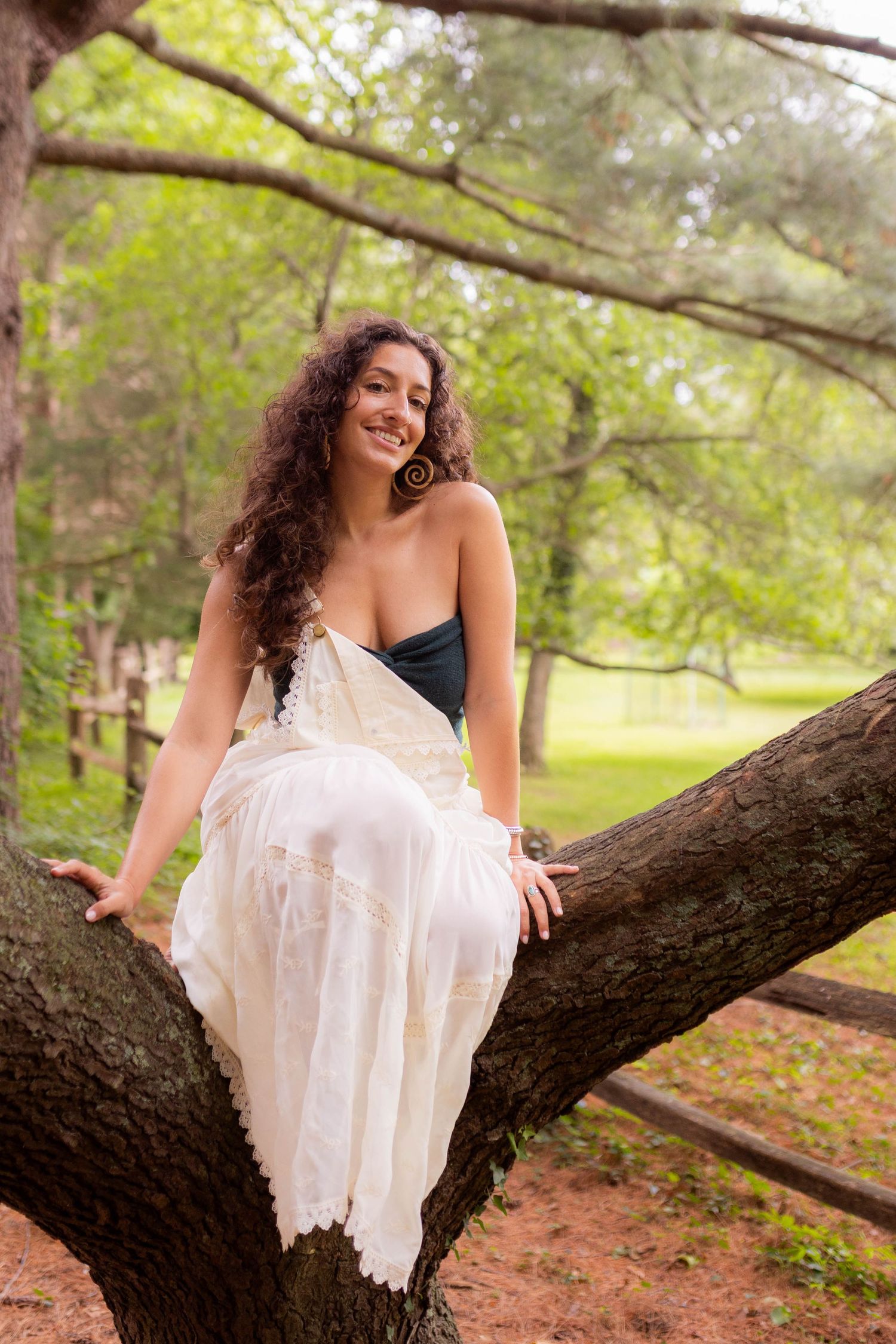 A person in a white dress sits on a tree branch in a sunny park setting.
