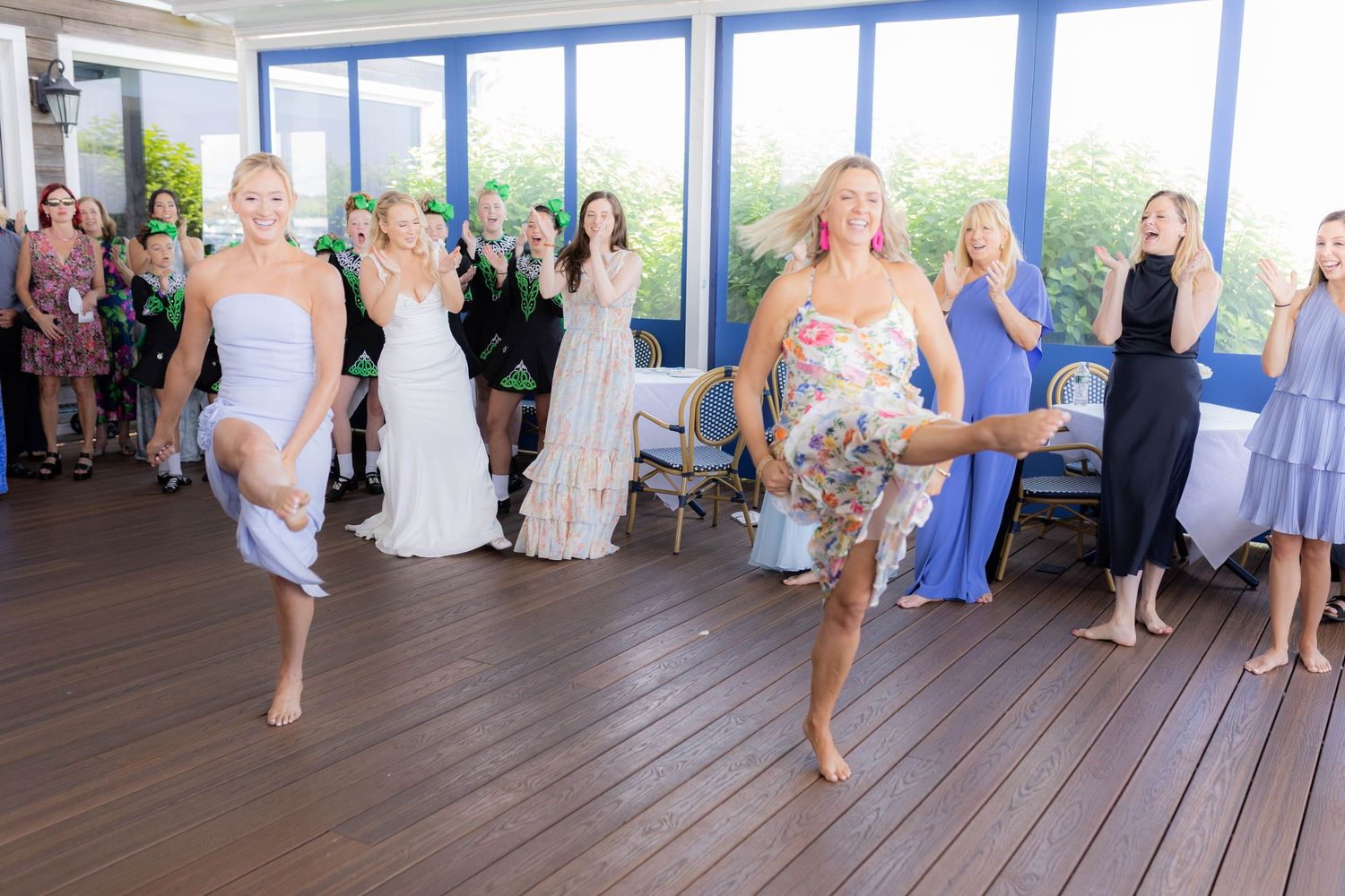 Wedding guests dancing energetically in blue and white dresses on a wooden deck.