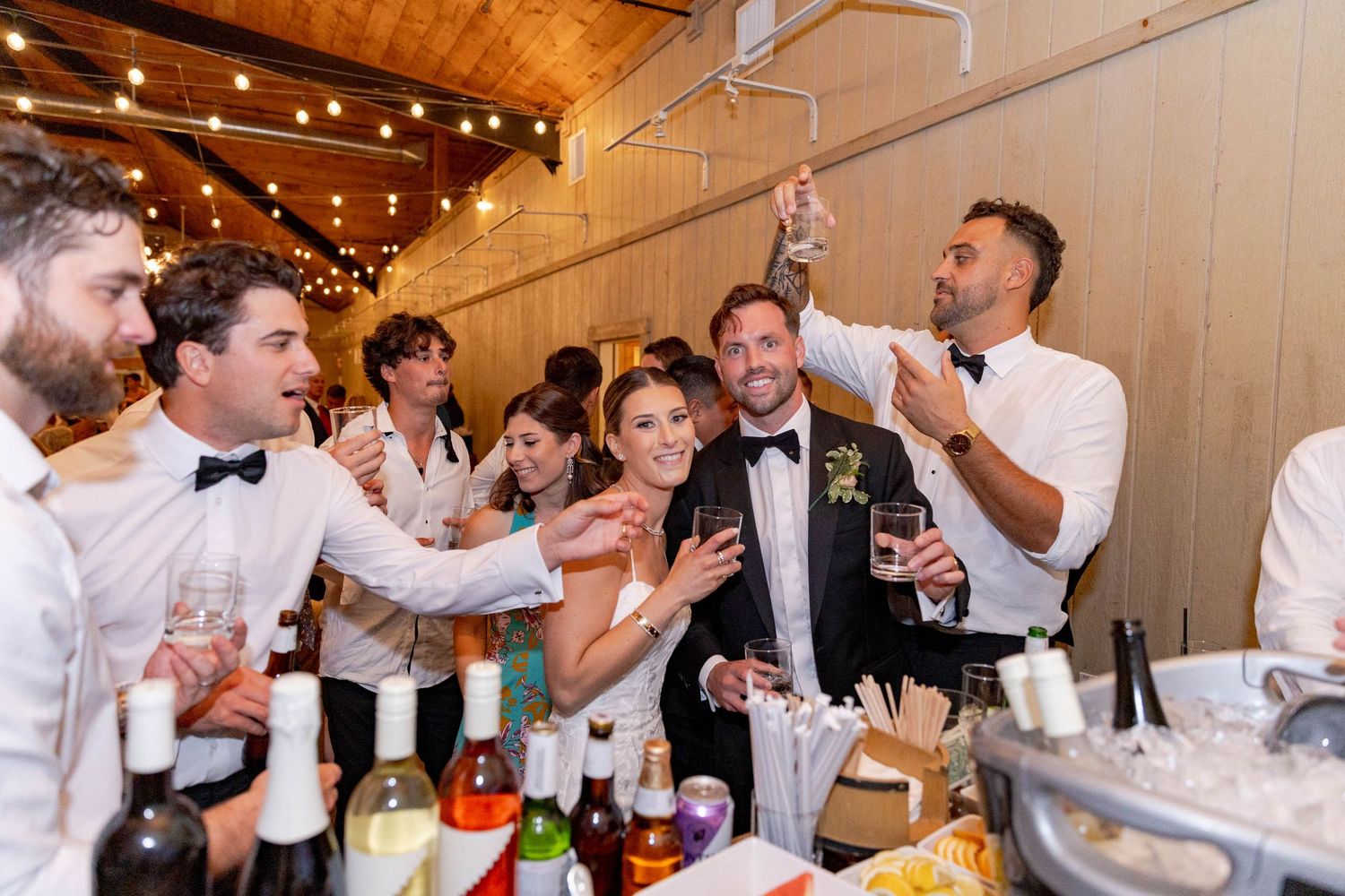 Wedding guests dancing and celebrating on a wooden dance floor under string lights in a rustic venue.