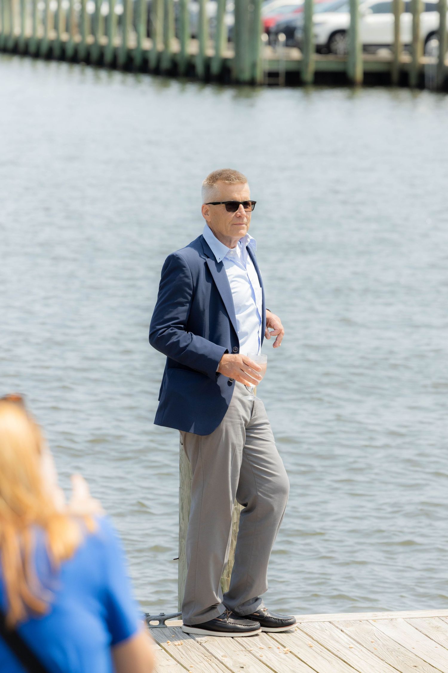 A person in a navy blazer and gray pants kneels down on a wooden dock by the water.