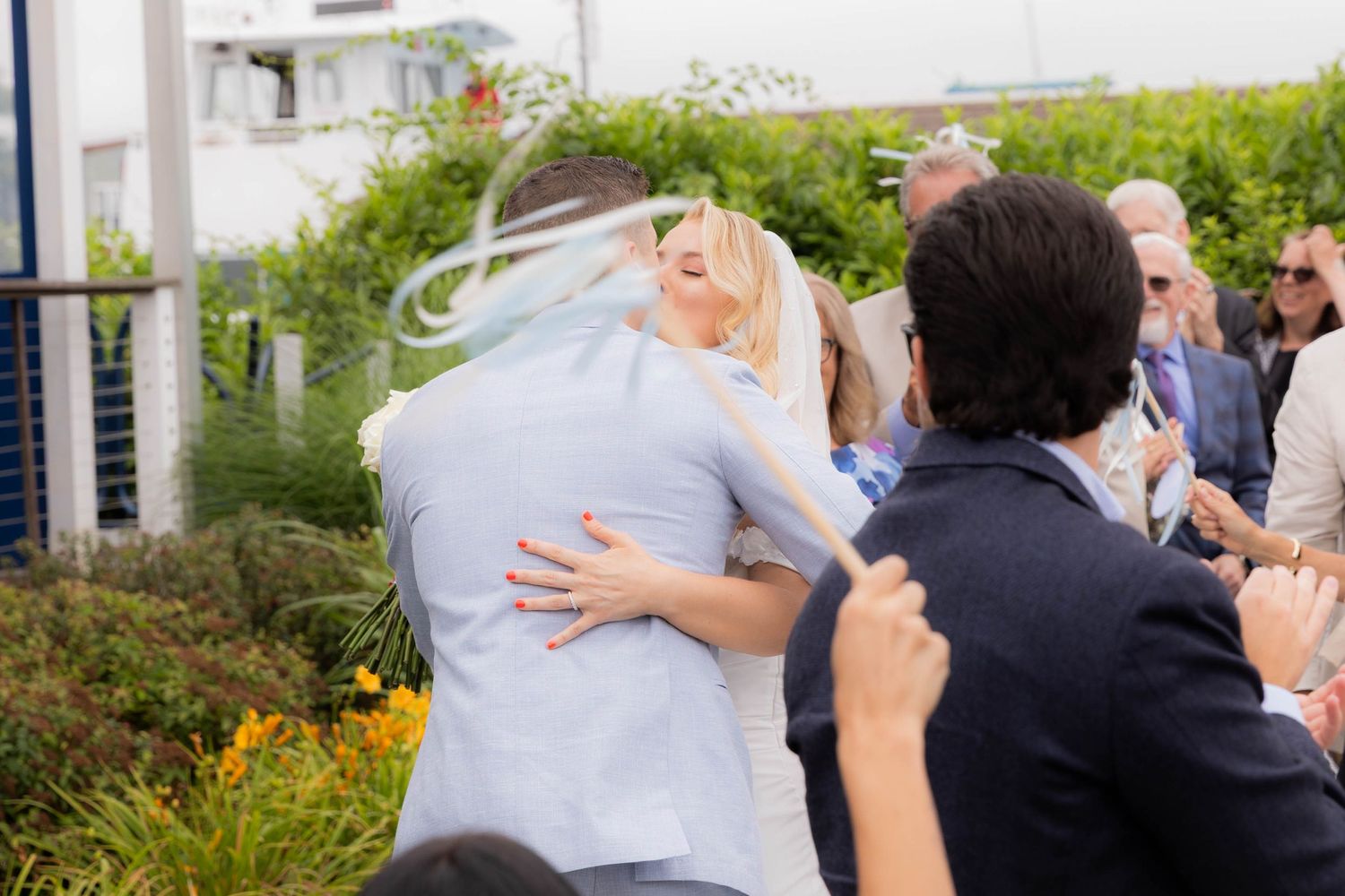 A joyful embrace between wedding guests during an outdoor celebration with greenery in the background.