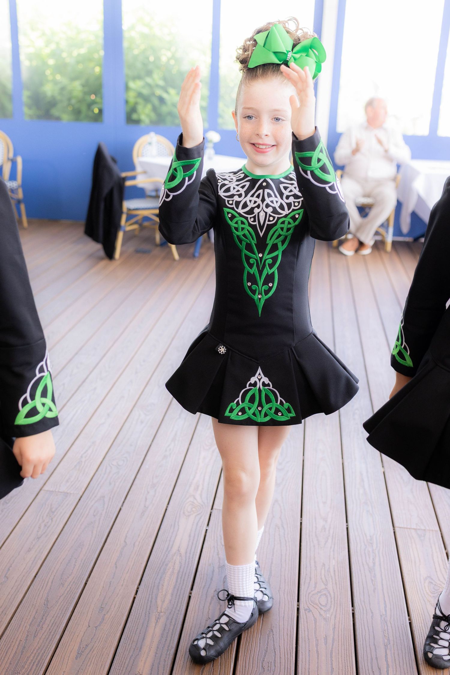 Irish dancer in black and green dress performs traditional steps on wooden deck during outdoor performance.