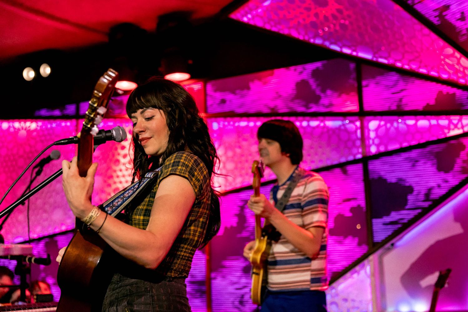 Band performs under intense pink stage lighting with geometric ceiling patterns.