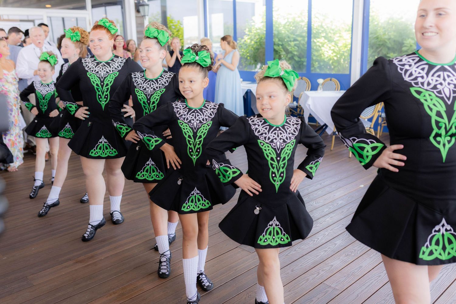 Irish dancers in black and green costumes perform traditional dance steps on a wooden deck.