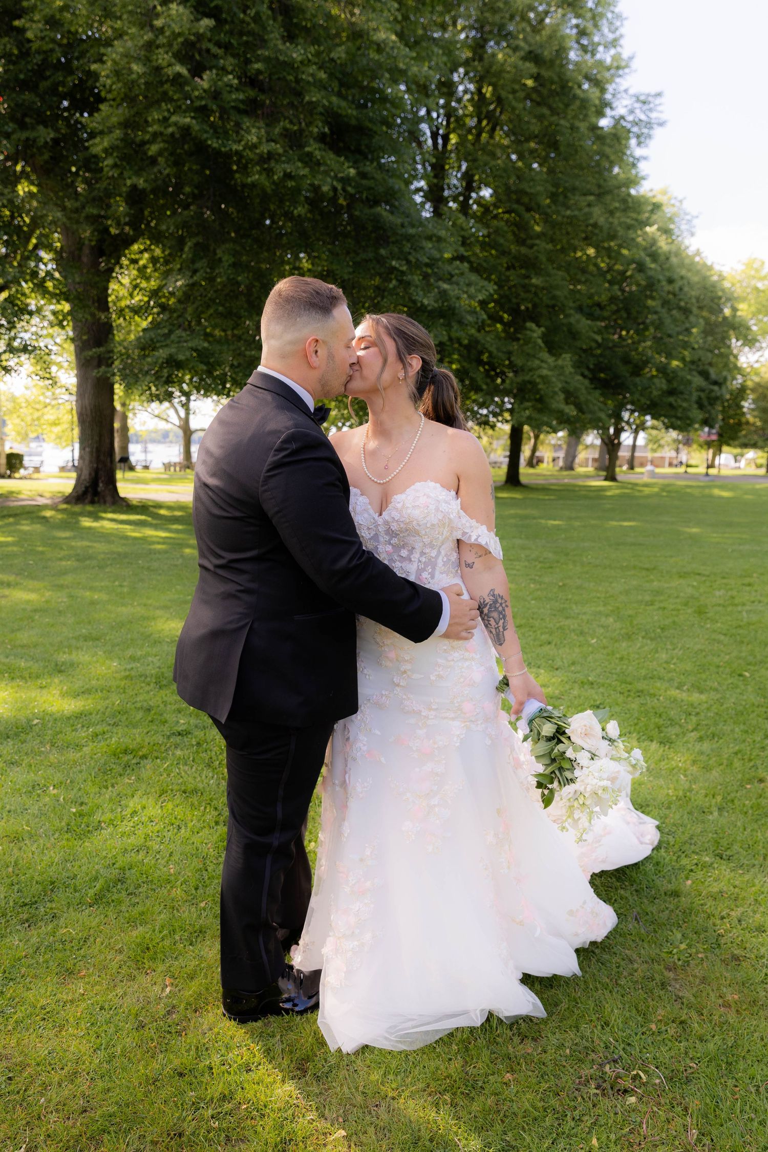 A couple shares a romantic kiss on their wedding day in a sunlit park with lush green trees in the background.