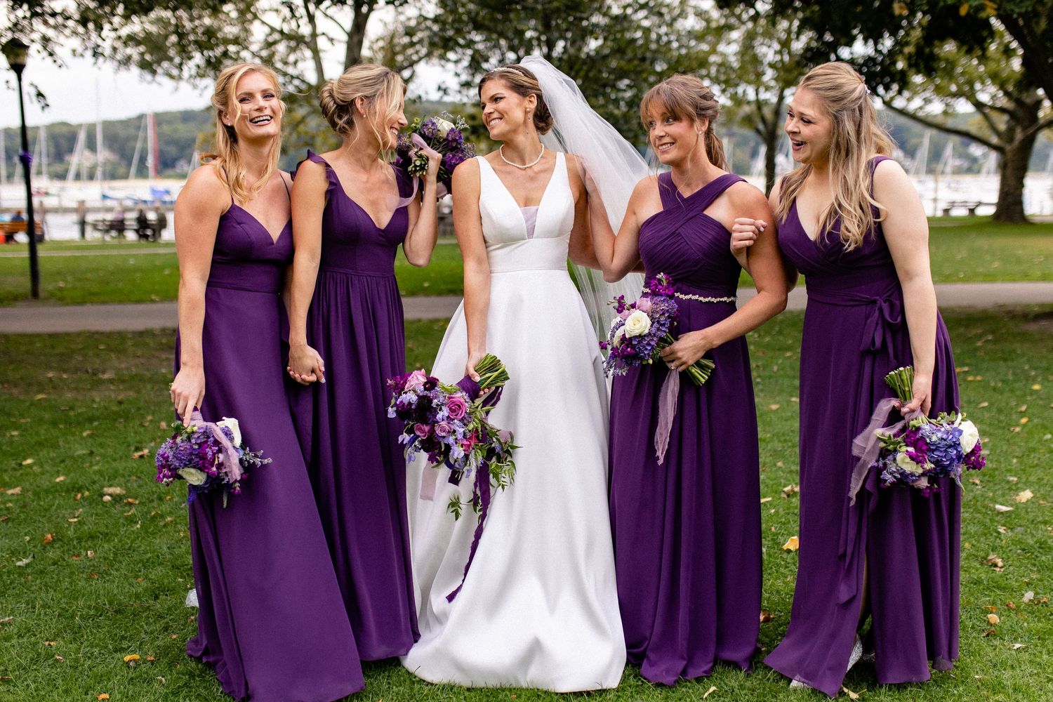 Wedding party in purple bridesmaid dresses and white bridal gown pose together outdoors by trees and water.