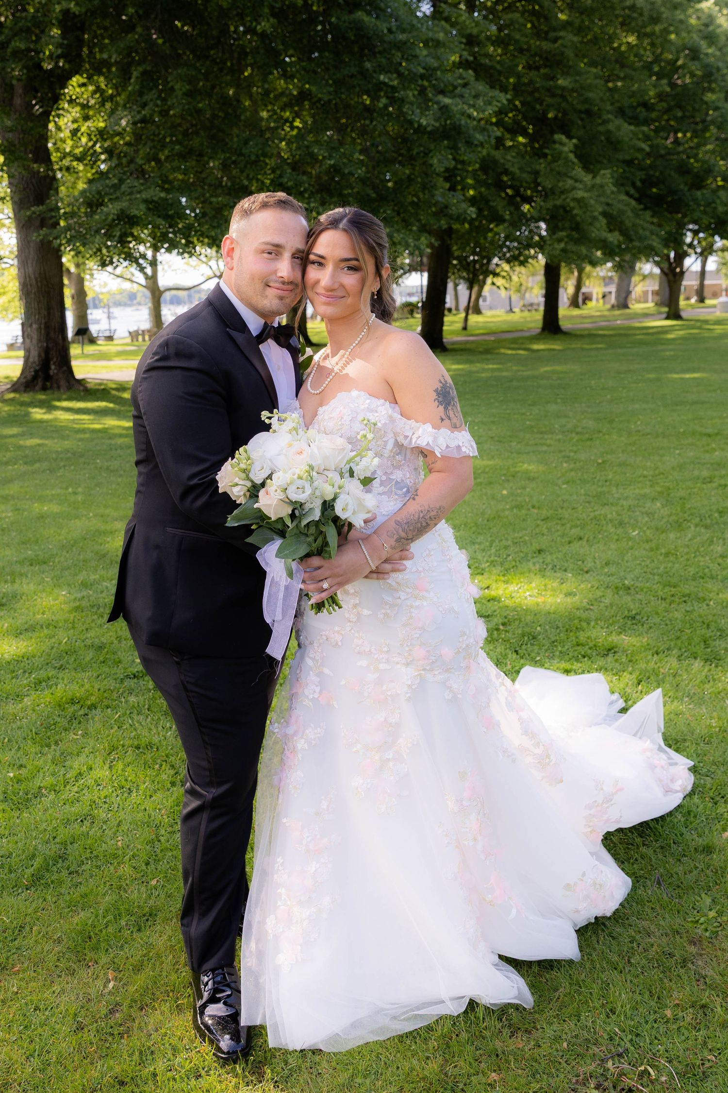 A bride in a lace off-shoulder gown and groom in black tuxedo pose together on a sunny lawn with white bouquet.
