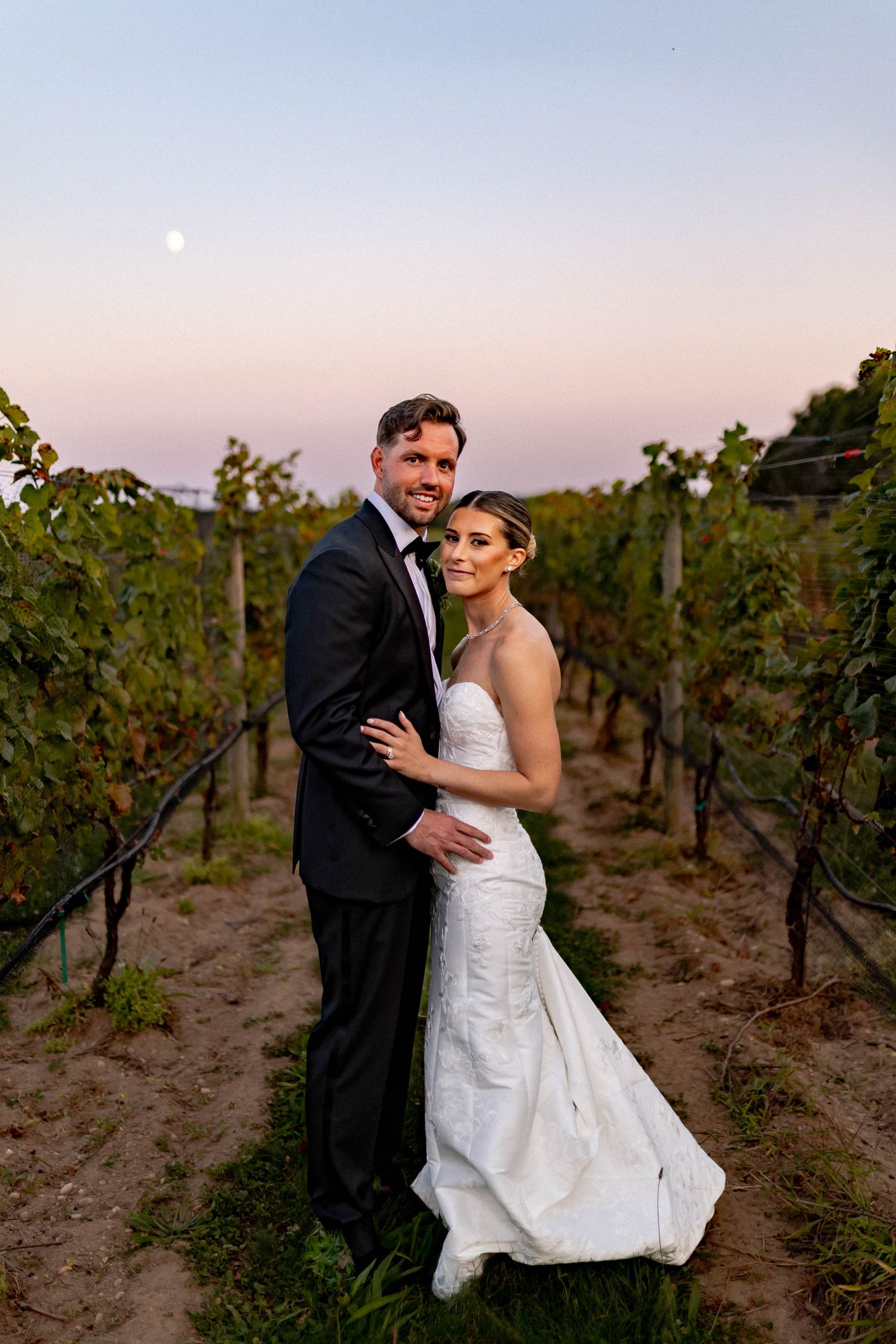 A couple embraces romantically at sunset among rows of grapevines in a vineyard.