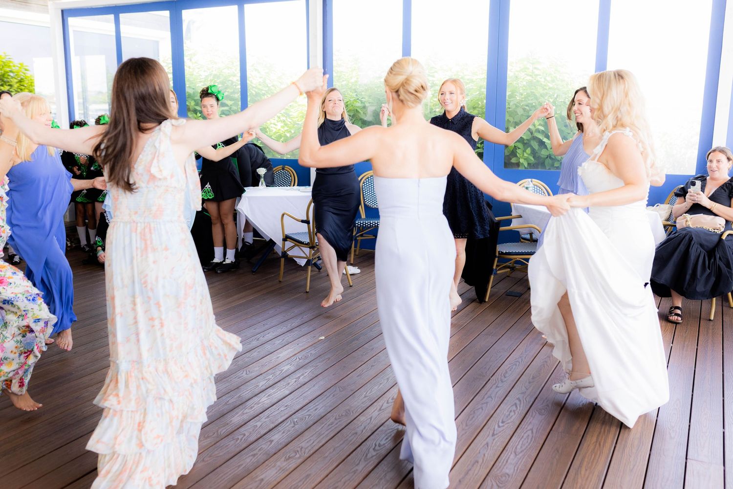 Wedding guests in white dresses dance together in a bright sunlit room with wooden floors.