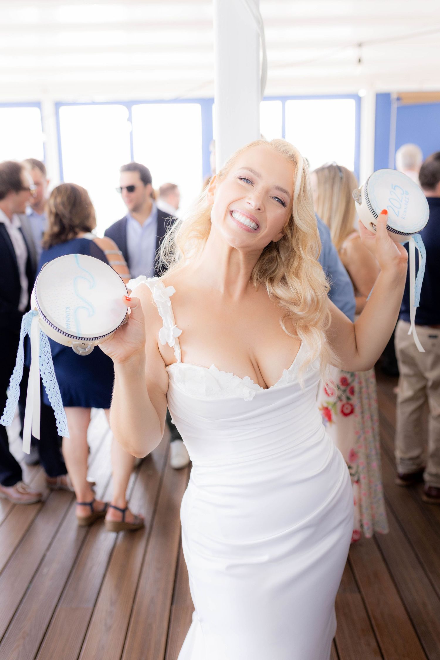 A bride in a white dress dances joyfully on a wooden deck with guests visible in the background.