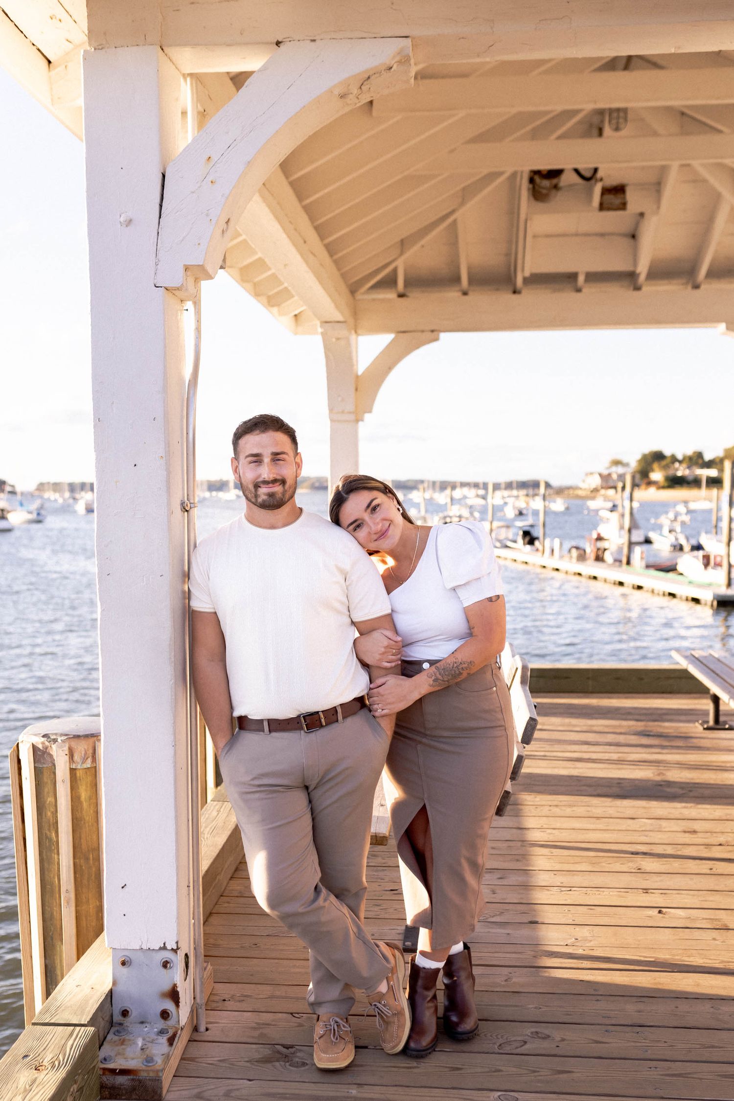 A couple poses together on a wooden pier with white columns and water views in the background.