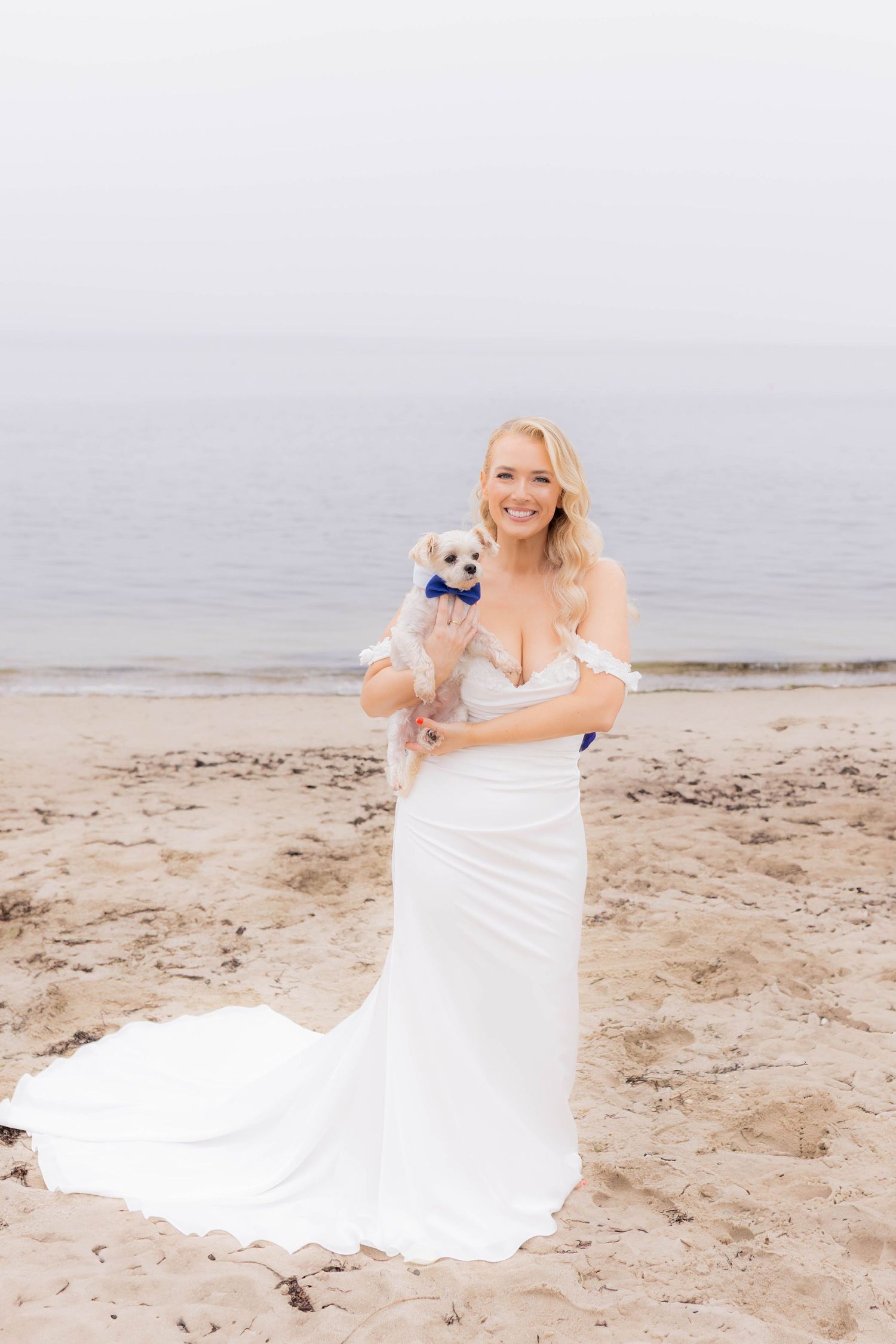 A white wedding dress flows in the breeze as a small white dog joins for beach photos.