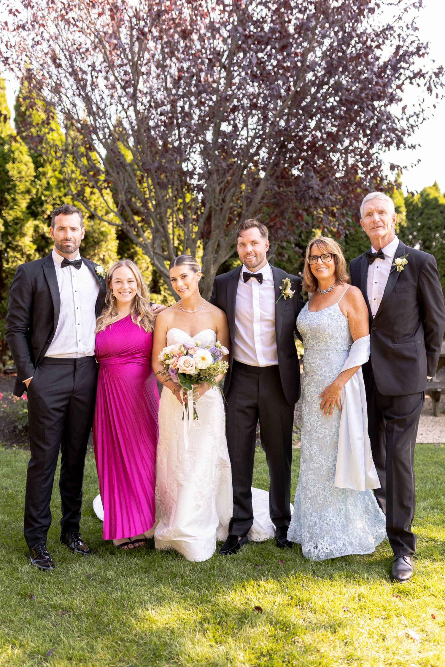 Wedding party in formal attire poses outdoors with bridesmaids in pink and light blue dresses.