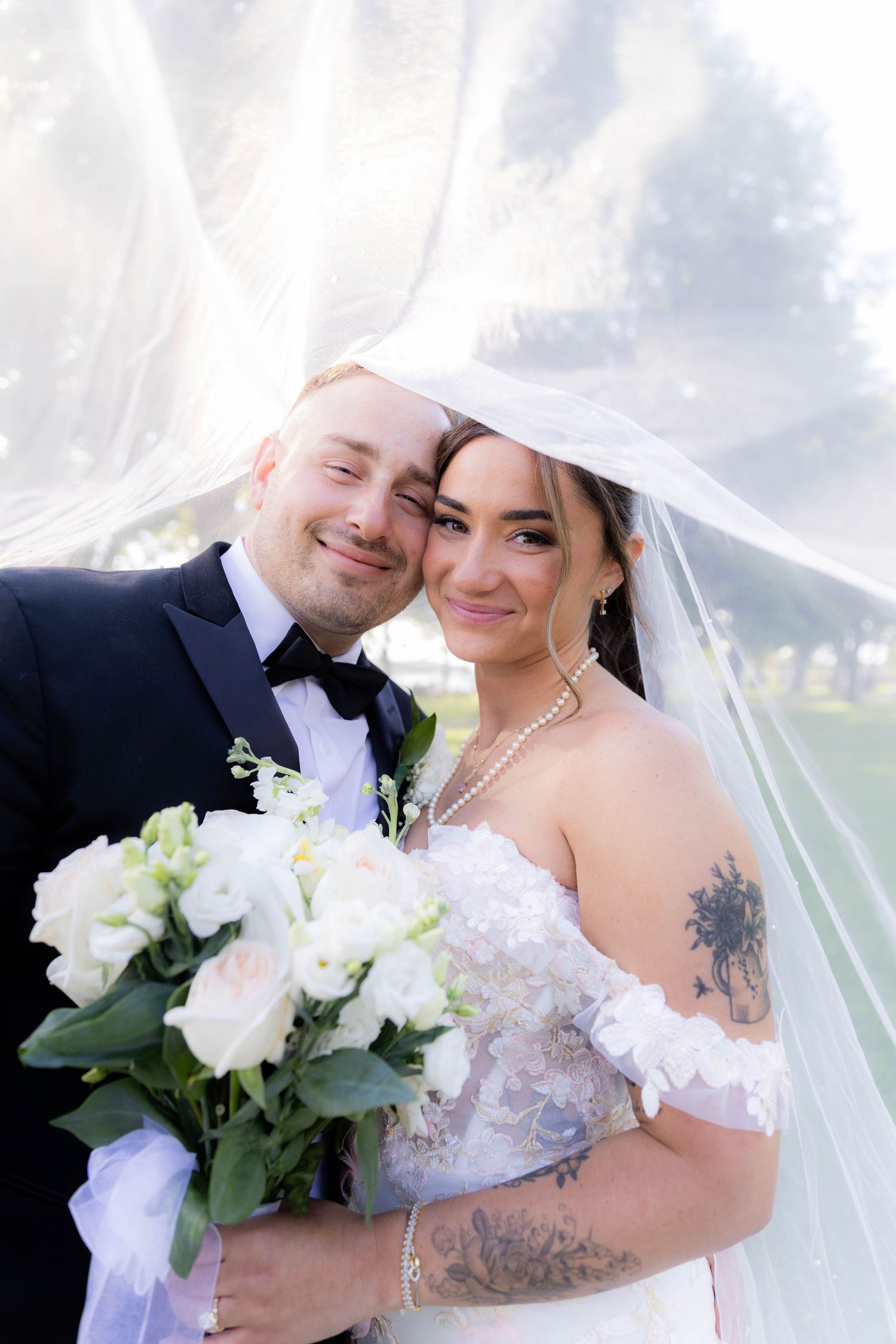A joyful wedding couple poses together under a flowing white veil with a bouquet of white flowers.