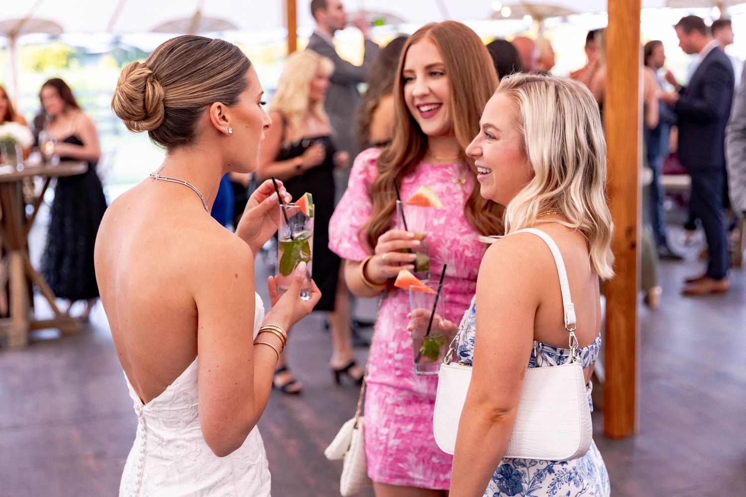 A group of wedding guests in pink and white dresses laugh and dance together at an outdoor reception.