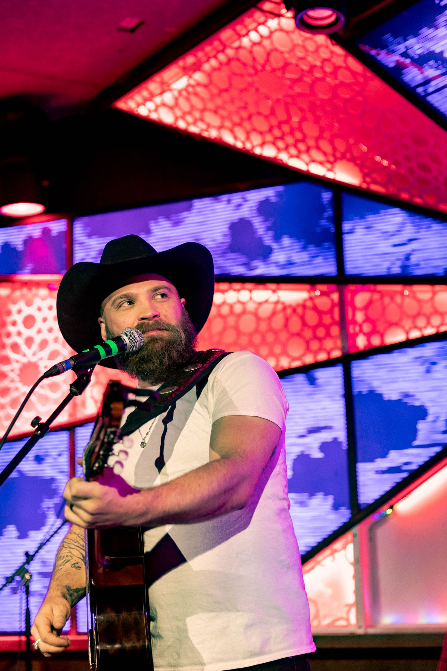 Country performer in white shirt and black hat sings into microphone on colorfully lit stage.