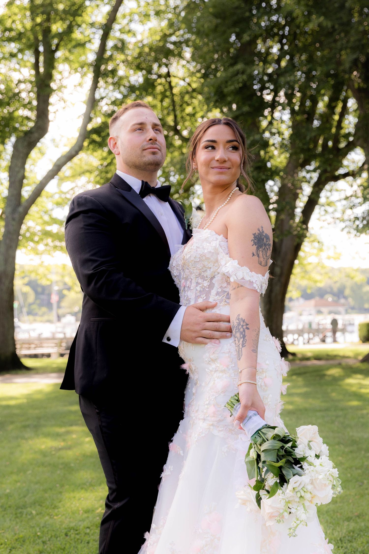 Romantic wedding couple embraces in sunlit garden setting with lush trees and greenery in the background.
