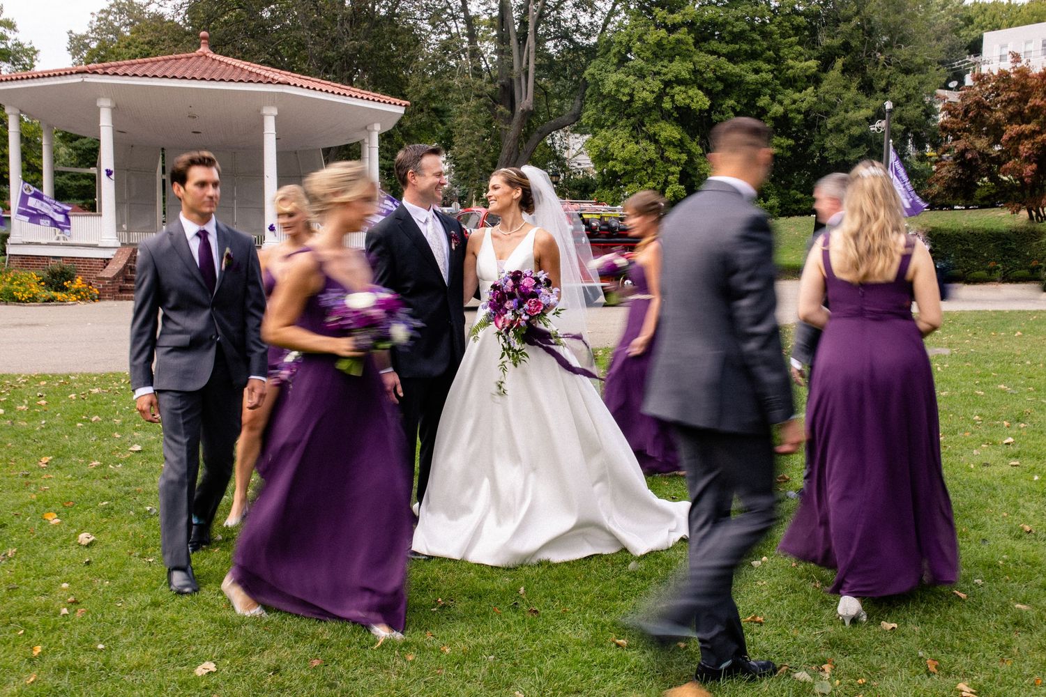 Wedding party members in purple dresses and dark suits playfully run across a green lawn during outdoor photo session.