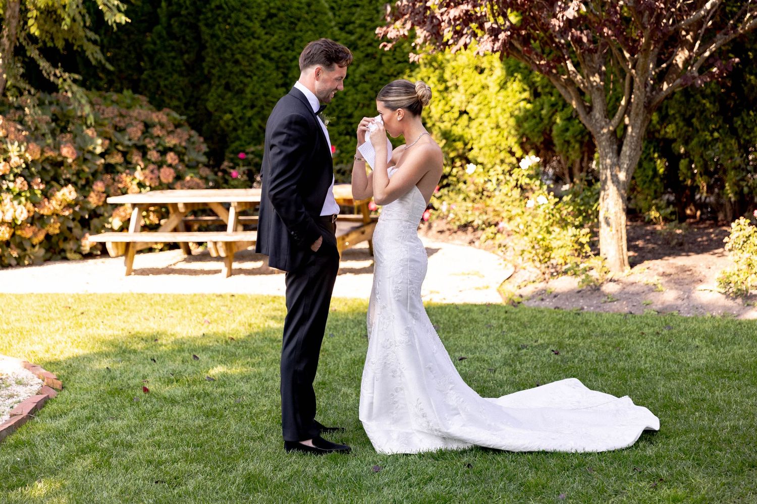 A couple in formal wedding attire shares a romantic moment in a sunlit garden setting.