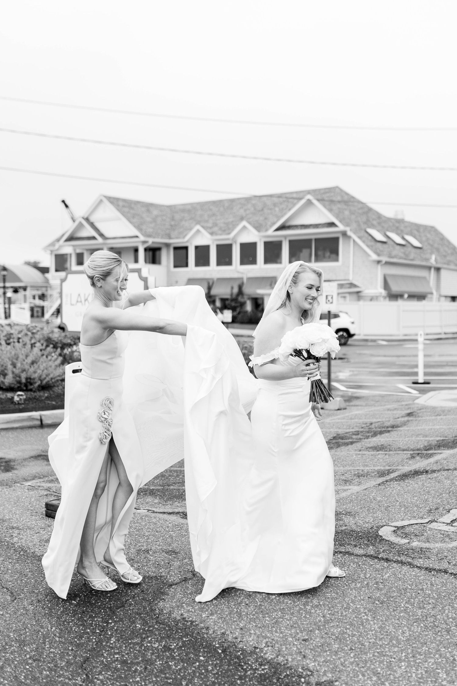 A black and white wedding photo showing a bride in a flowing dress with someone helping adjust her veil outdoors.