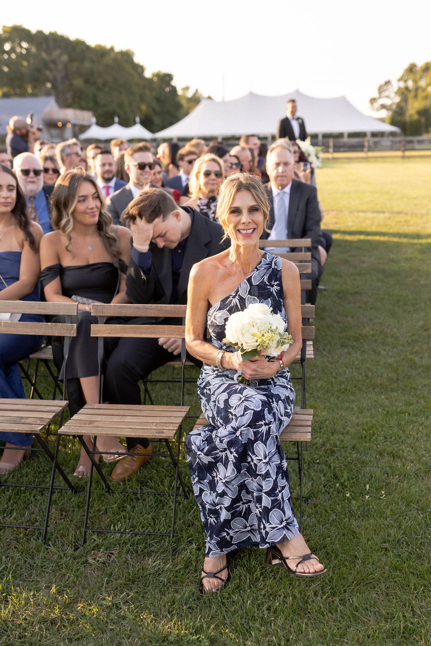 Wedding guests seated on wooden benches in a grassy field during an outdoor ceremony.