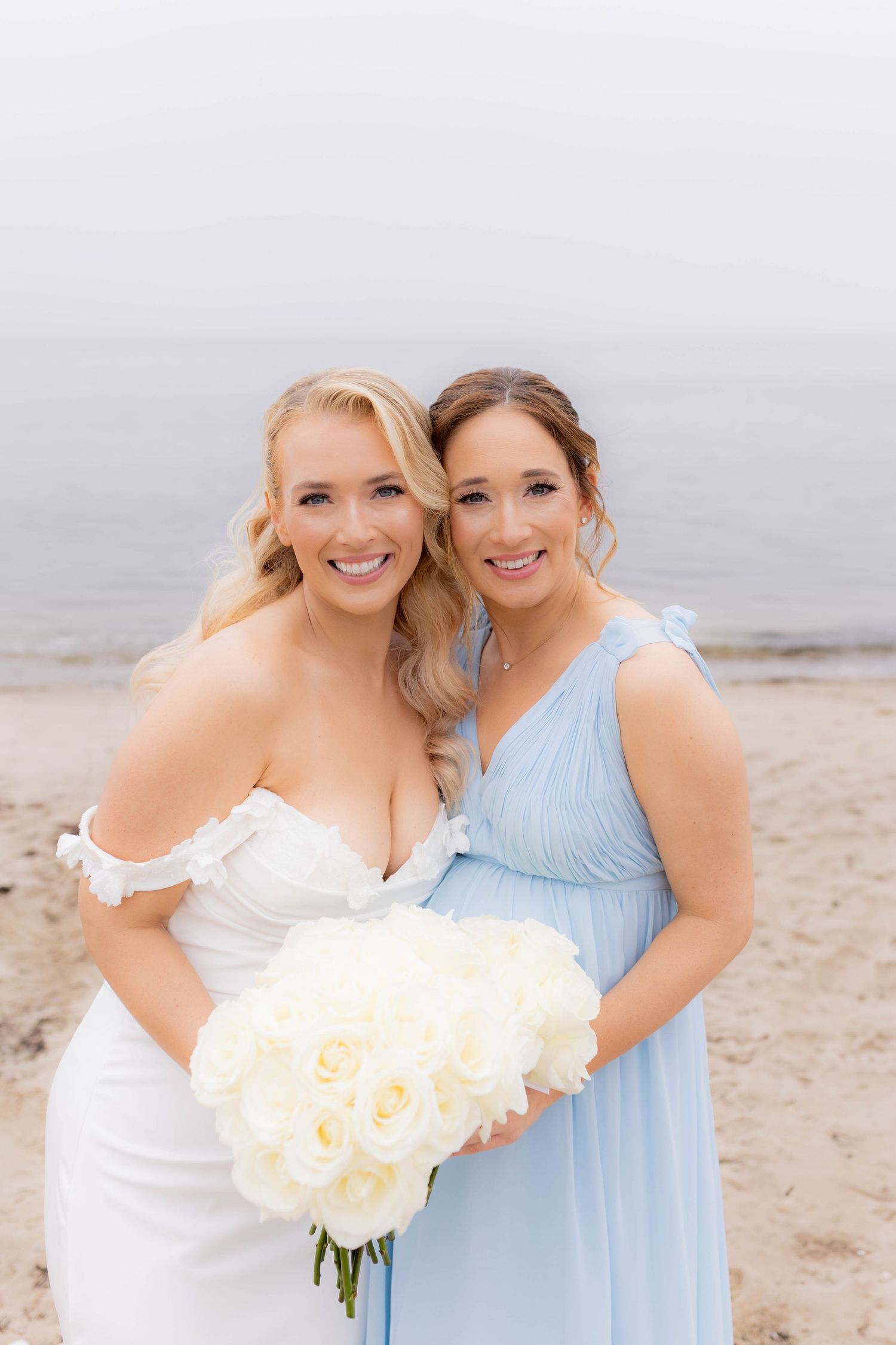 Two women in light blue and white dresses pose together on a beach holding floral bouquets.