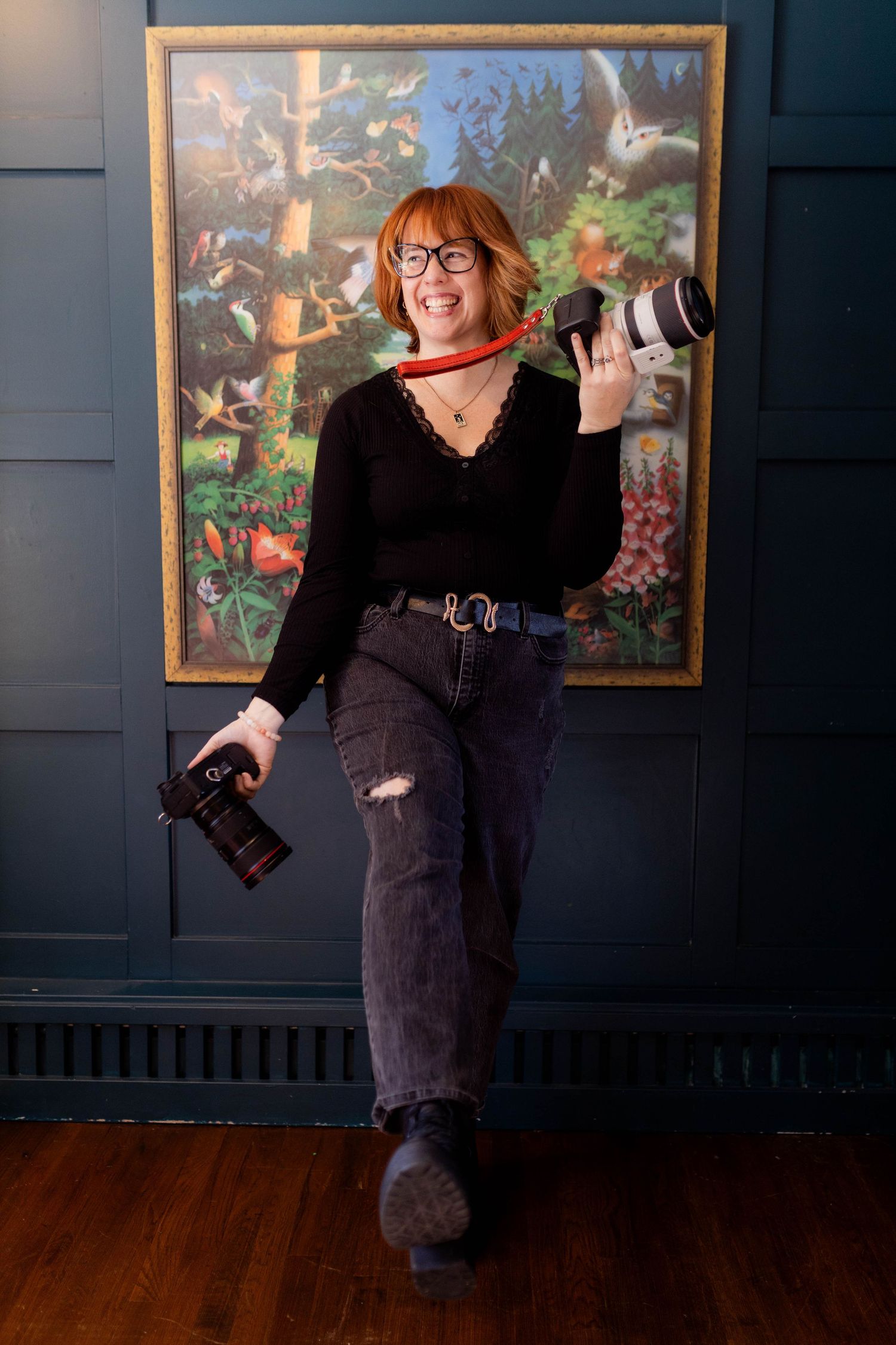 Photographer poses with camera against dark wall with colorful framed artwork. Stephanie Coiro. Fireside Sparrow Photography