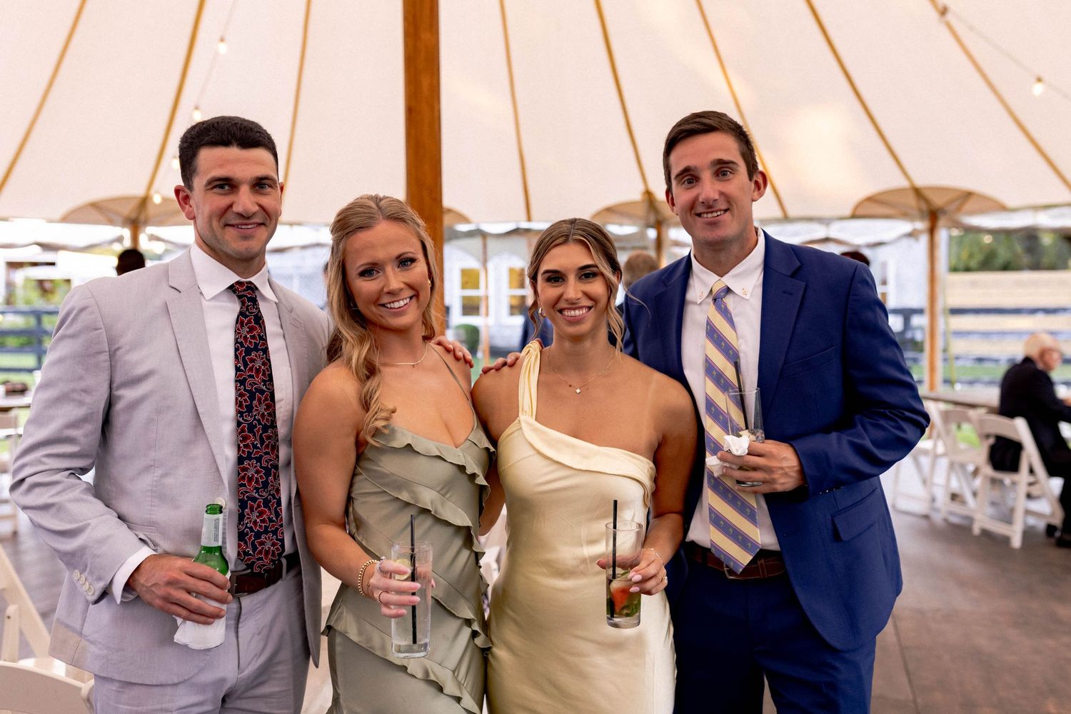 Group of wedding guests smiling and enjoying drinks together under a white tent at an outdoor reception.