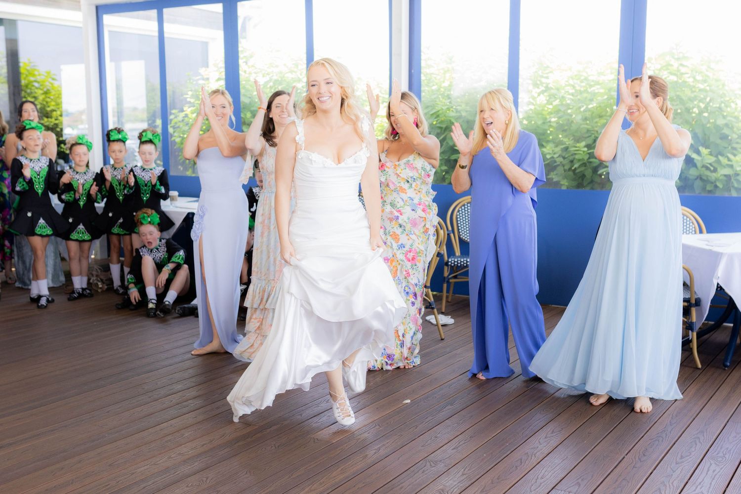 Wedding party members in blue dresses and white gowns dance together on a wooden deck during outdoor celebration.