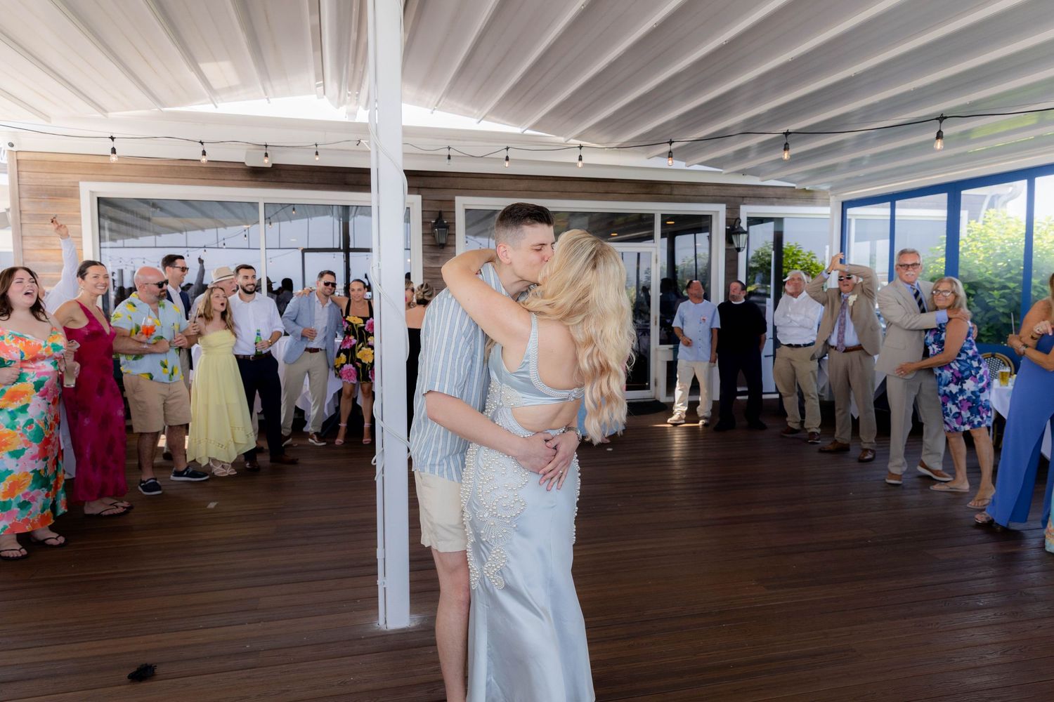 Wedding guests dancing and mingling in a bright reception venue with white beams and exposed ceiling.