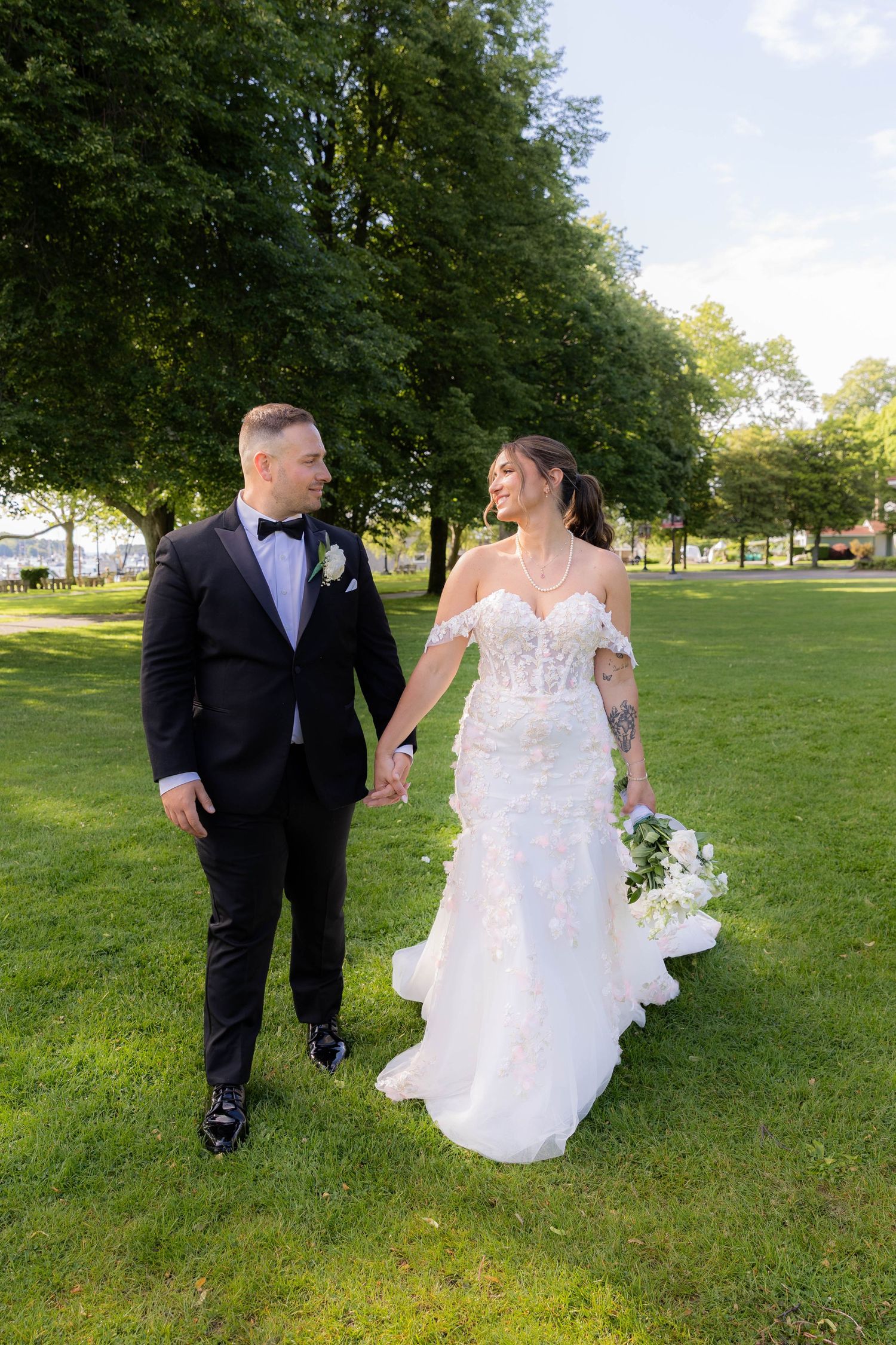 A couple in wedding attire walk hand in hand across a sunlit lawn surrounded by green trees.