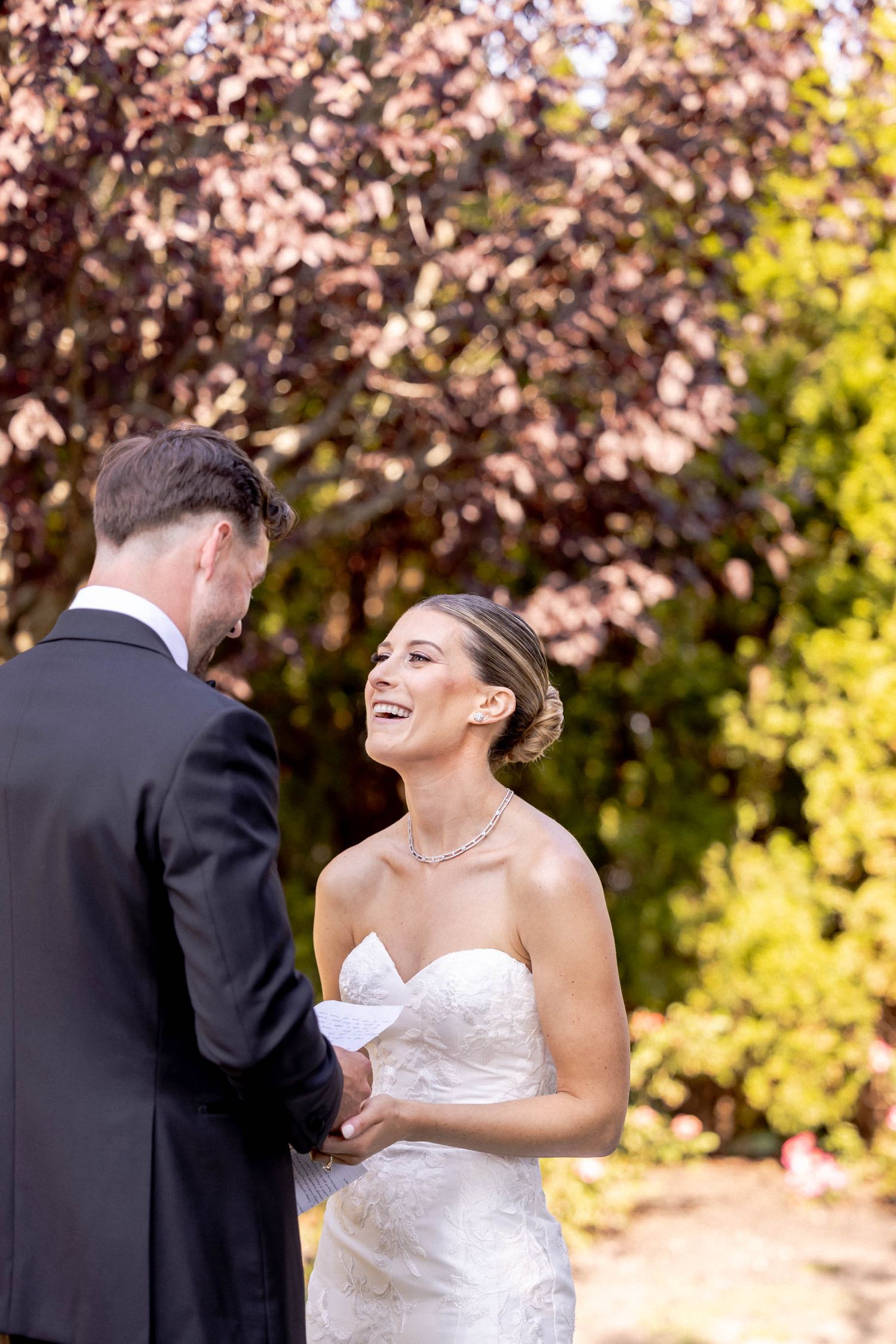 A bride in a white strapless gown laughs joyfully during an outdoor wedding ceremony with autumn foliage in background.