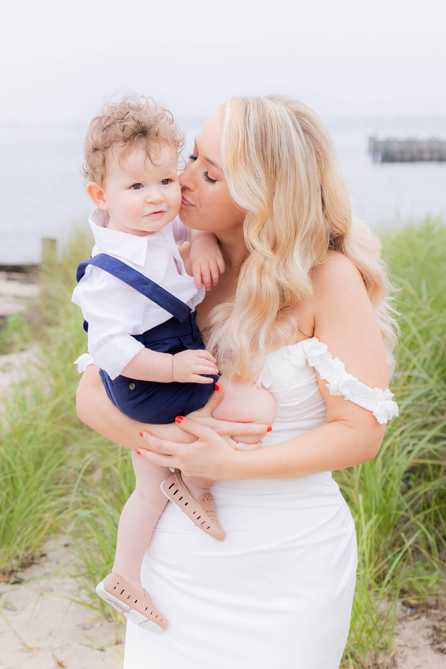 A tender moment captured at the beach with a woman in white dress holding a small child in navy blue outfit.