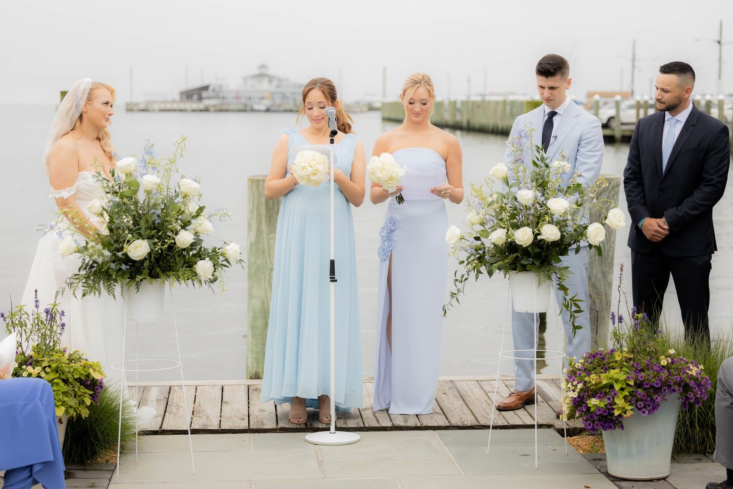 Wedding party in light blue dresses poses on a waterfront deck with floral bouquets.