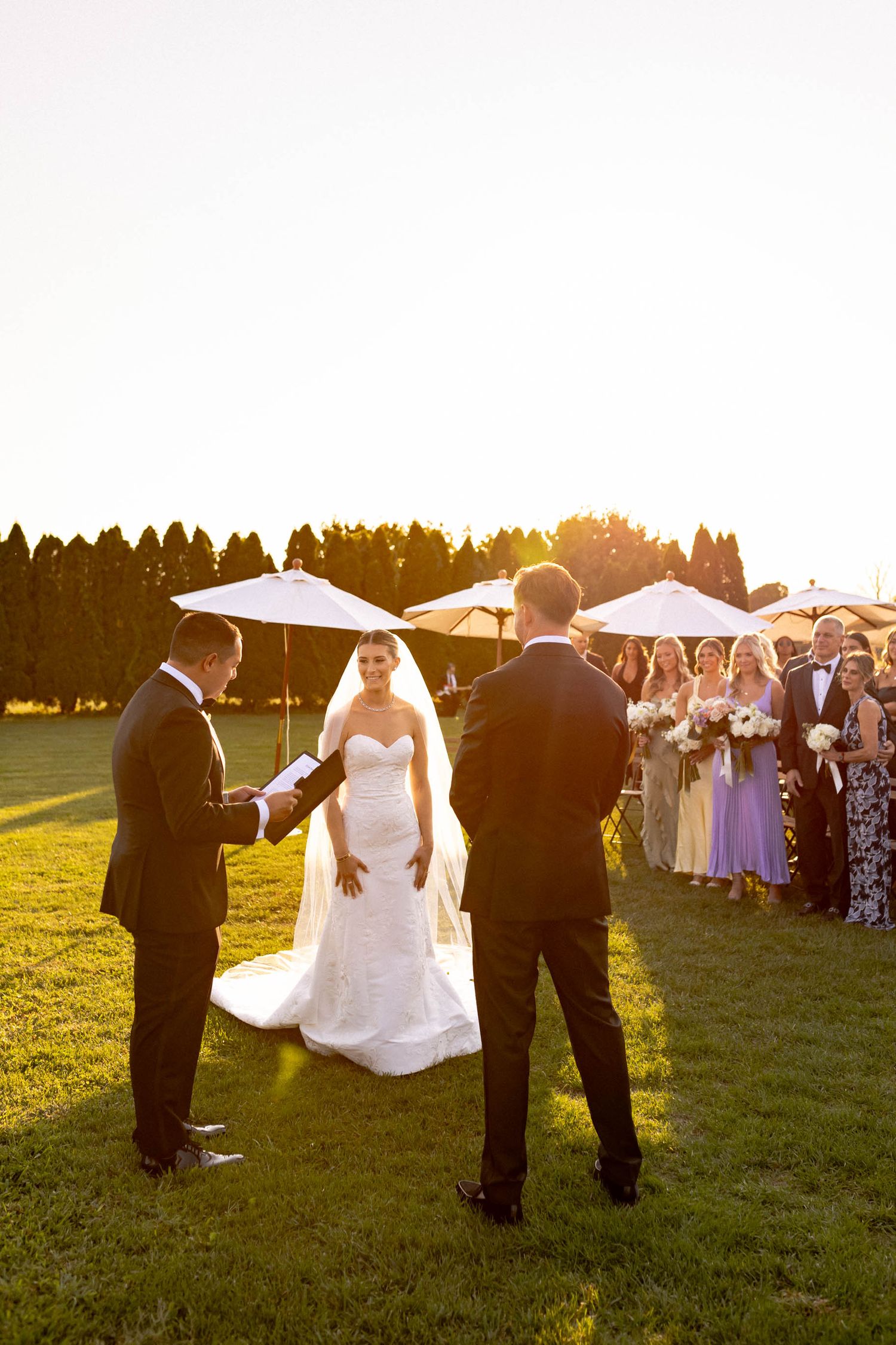 A sunset wedding ceremony takes place outdoors with attendants holding parasols on a grassy field.
