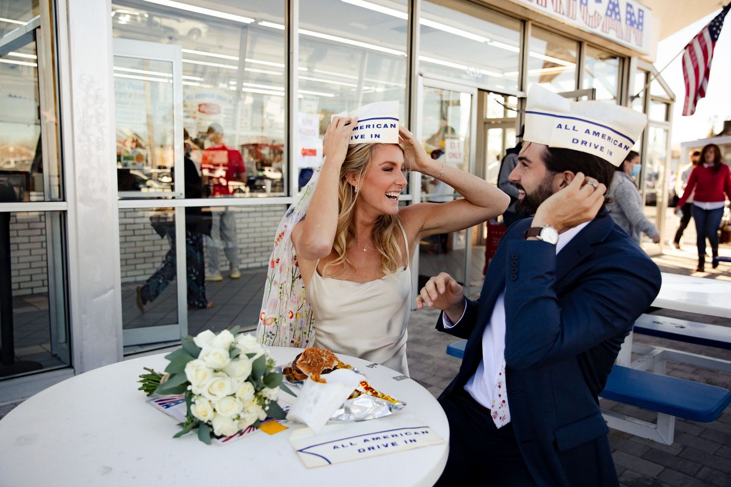 Newlyweds playfully sharing food at outdoor table with white floral bouquet. Fireside Sparrow Photography. All American Burger