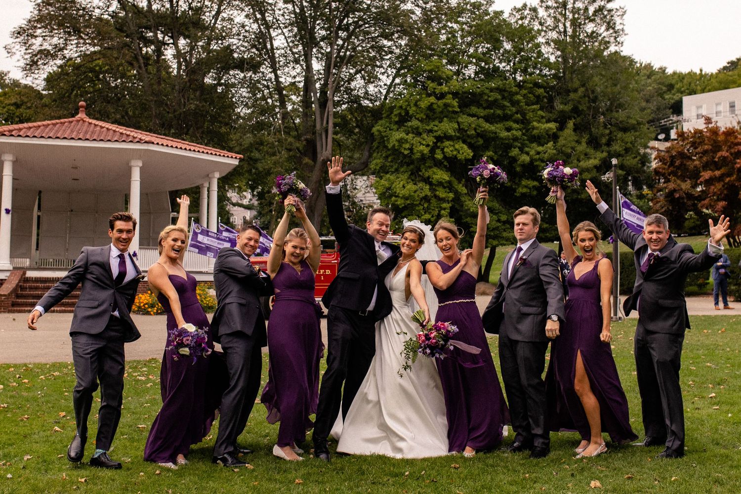 Wedding party in purple attire poses together on a grassy lawn during an outdoor celebration.