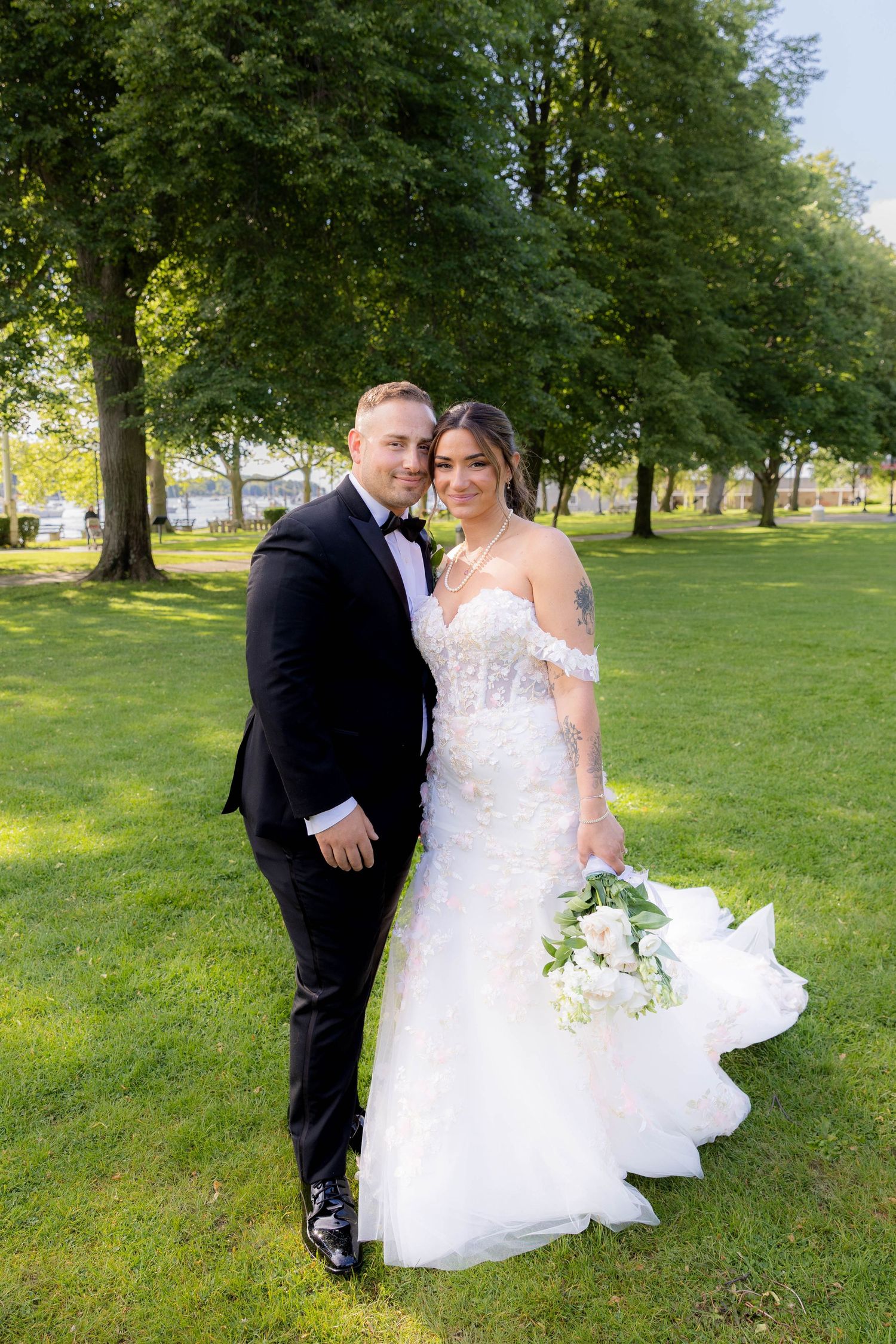 A couple in wedding attire poses together on a lush green lawn with trees in the background during a sunny day.