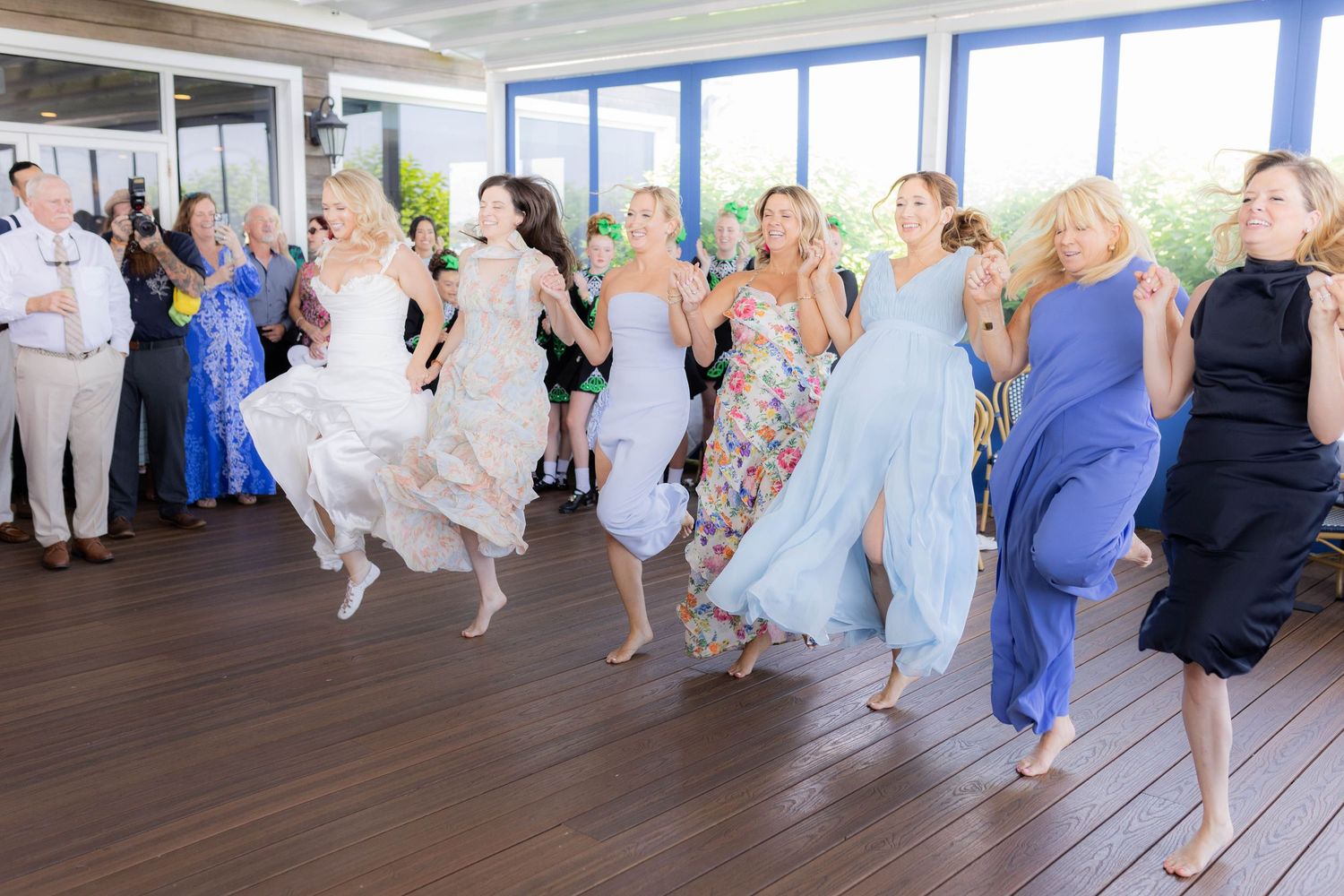 Group of wedding guests performing synchronized dance moves in pastel dresses on a wooden floor.