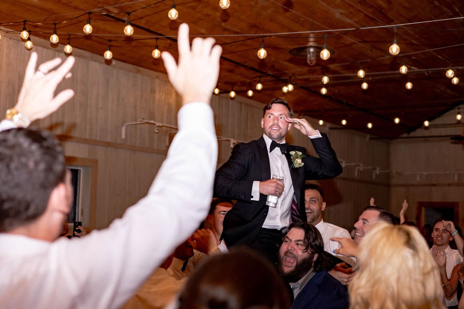 A crowd celebrates at a lively indoor party with string lights overhead and raised hands in celebration.