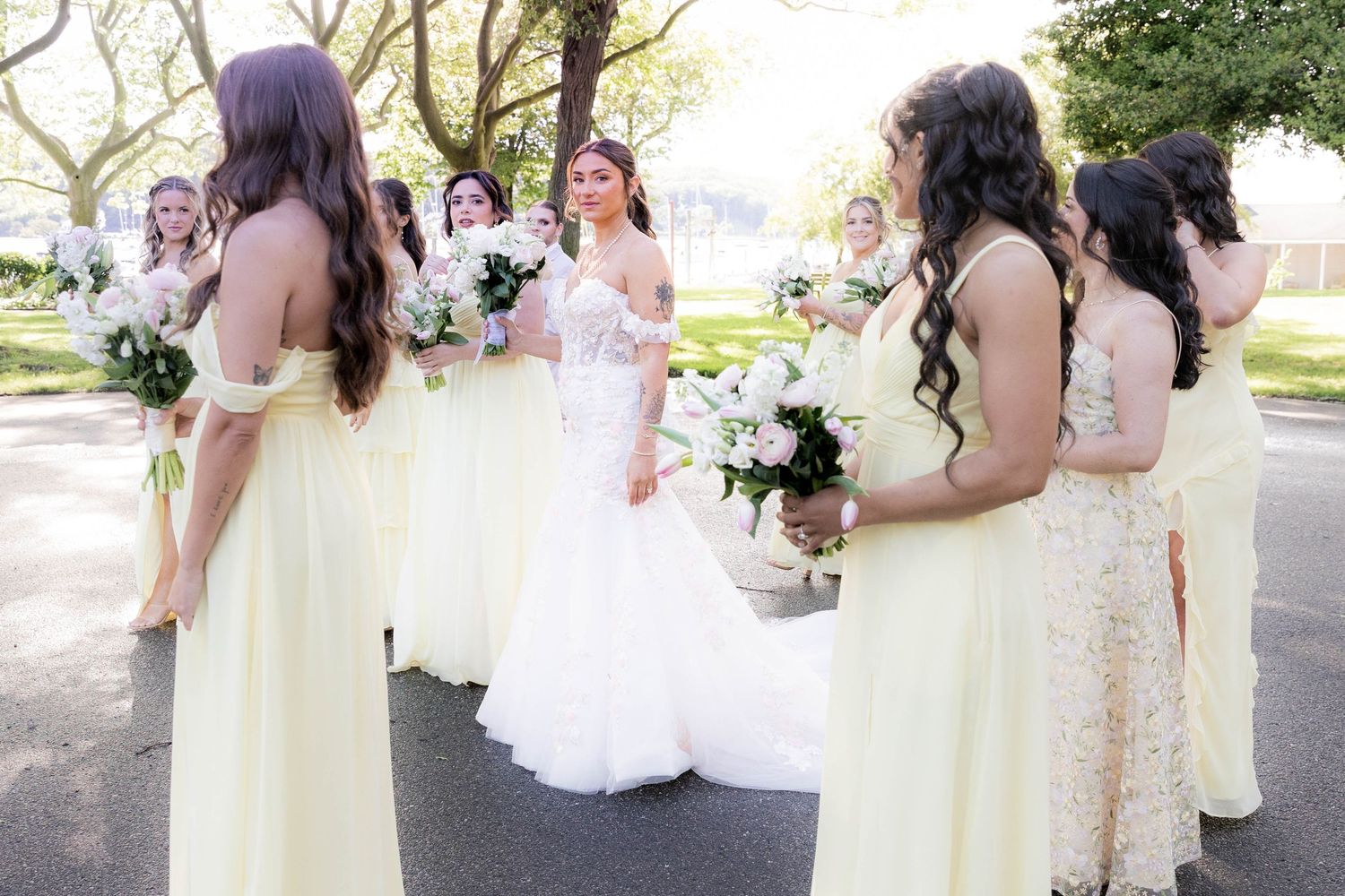 A group of bridesmaids in pastel yellow dresses pose together with bouquets at an outdoor wedding.
