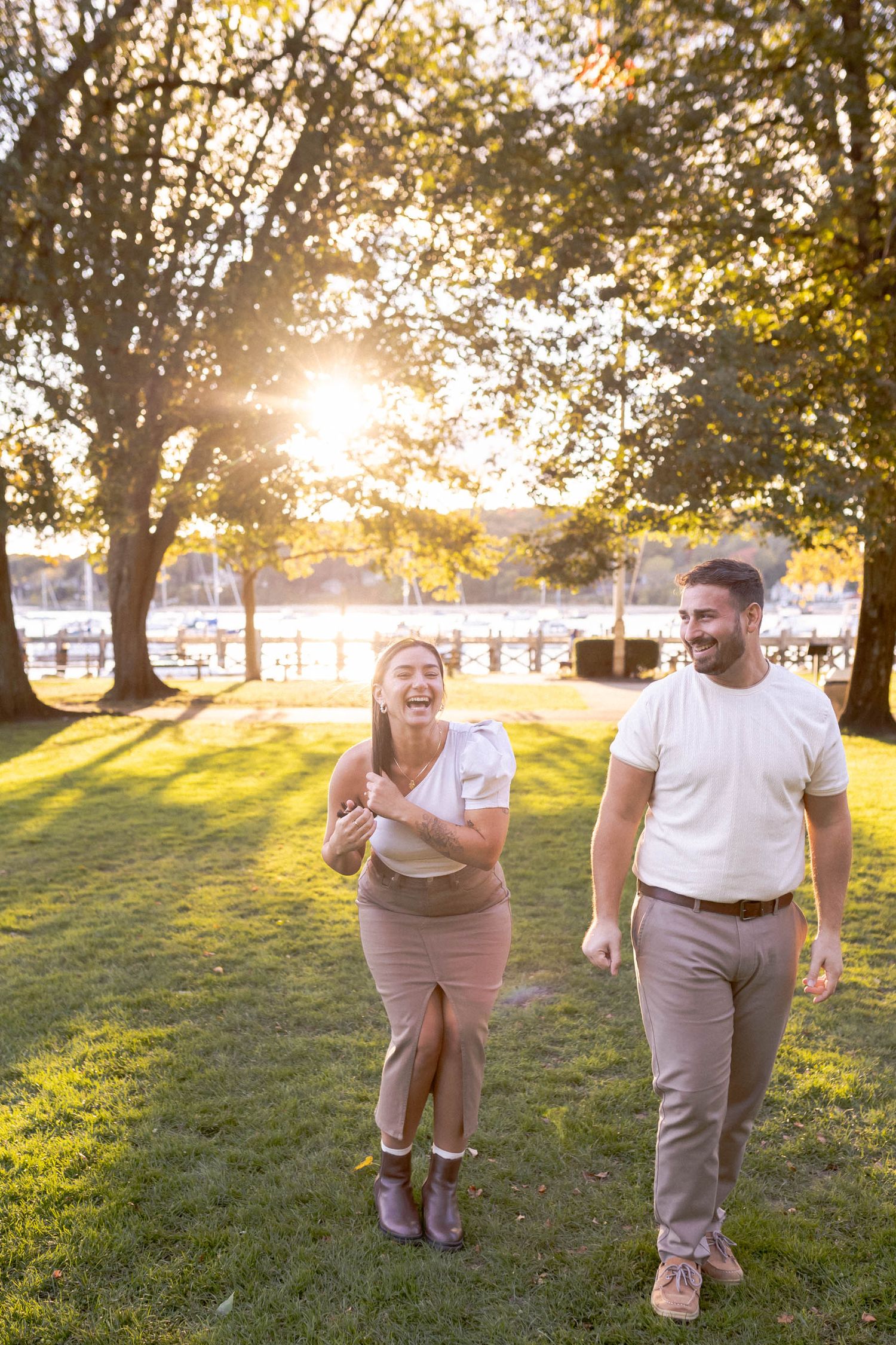 A couple enjoys a romantic sunset walk through a park with golden sunlight streaming through trees by the water.