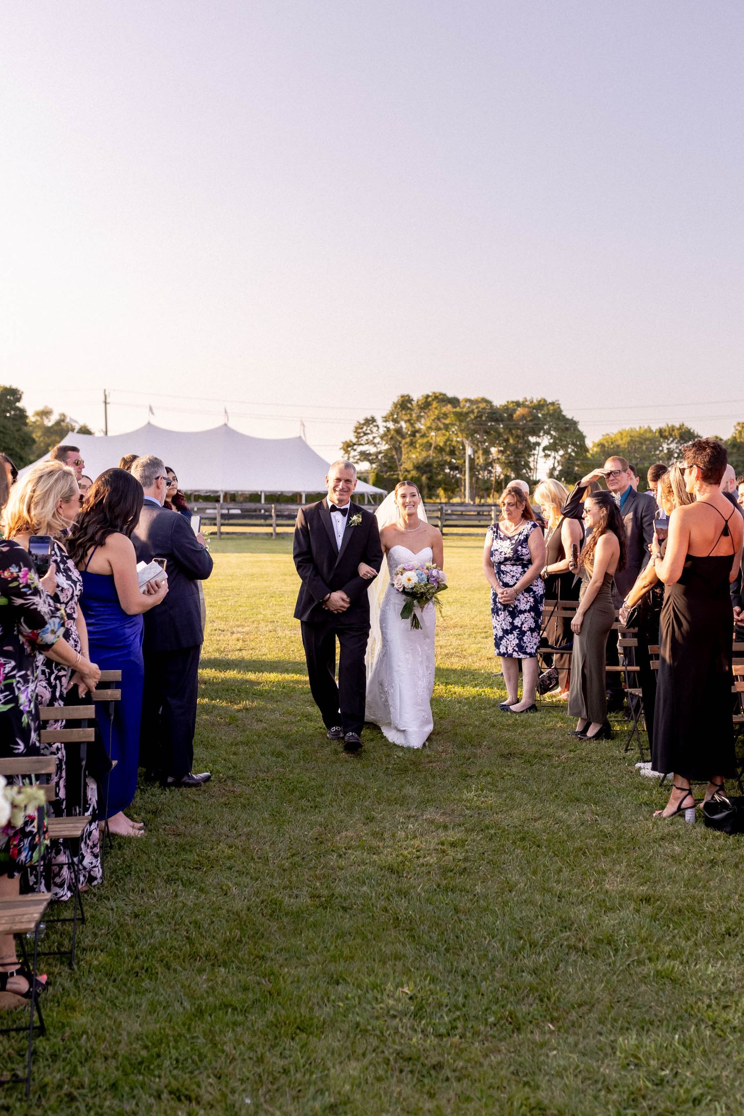 Wedding guests gathering on a lawn during an outdoor ceremony with a white tent visible.