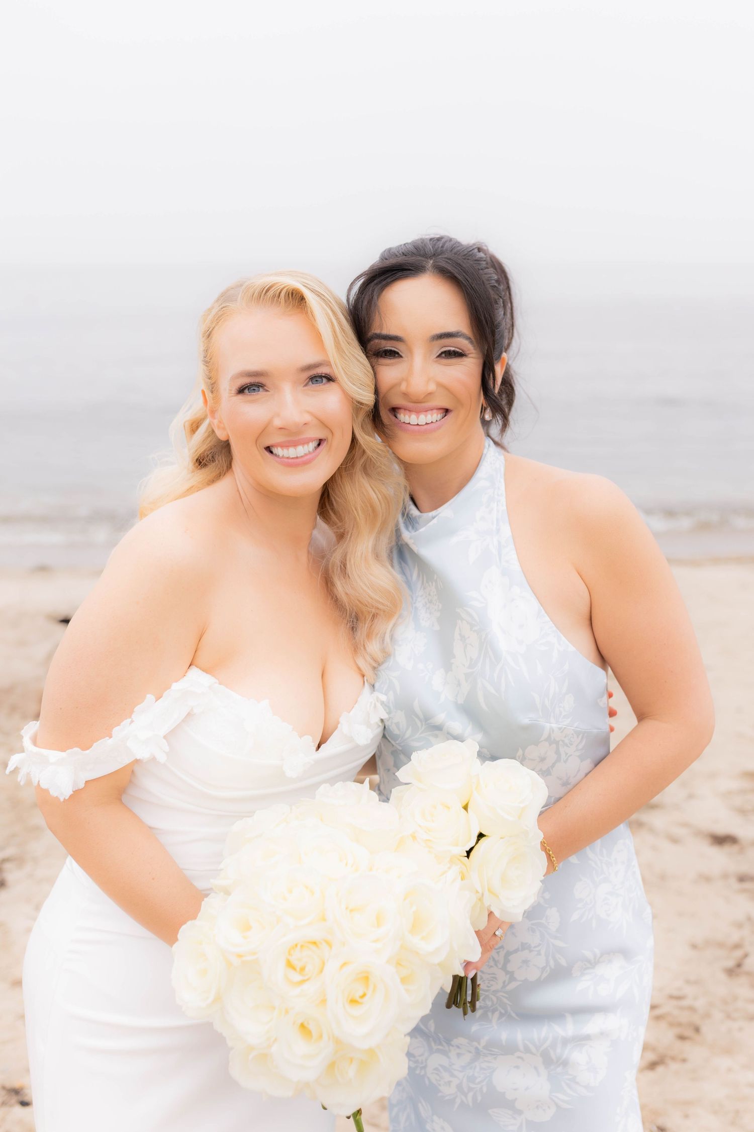 Two brides in white dresses smile together on a beach holding cream-colored rose bouquets.